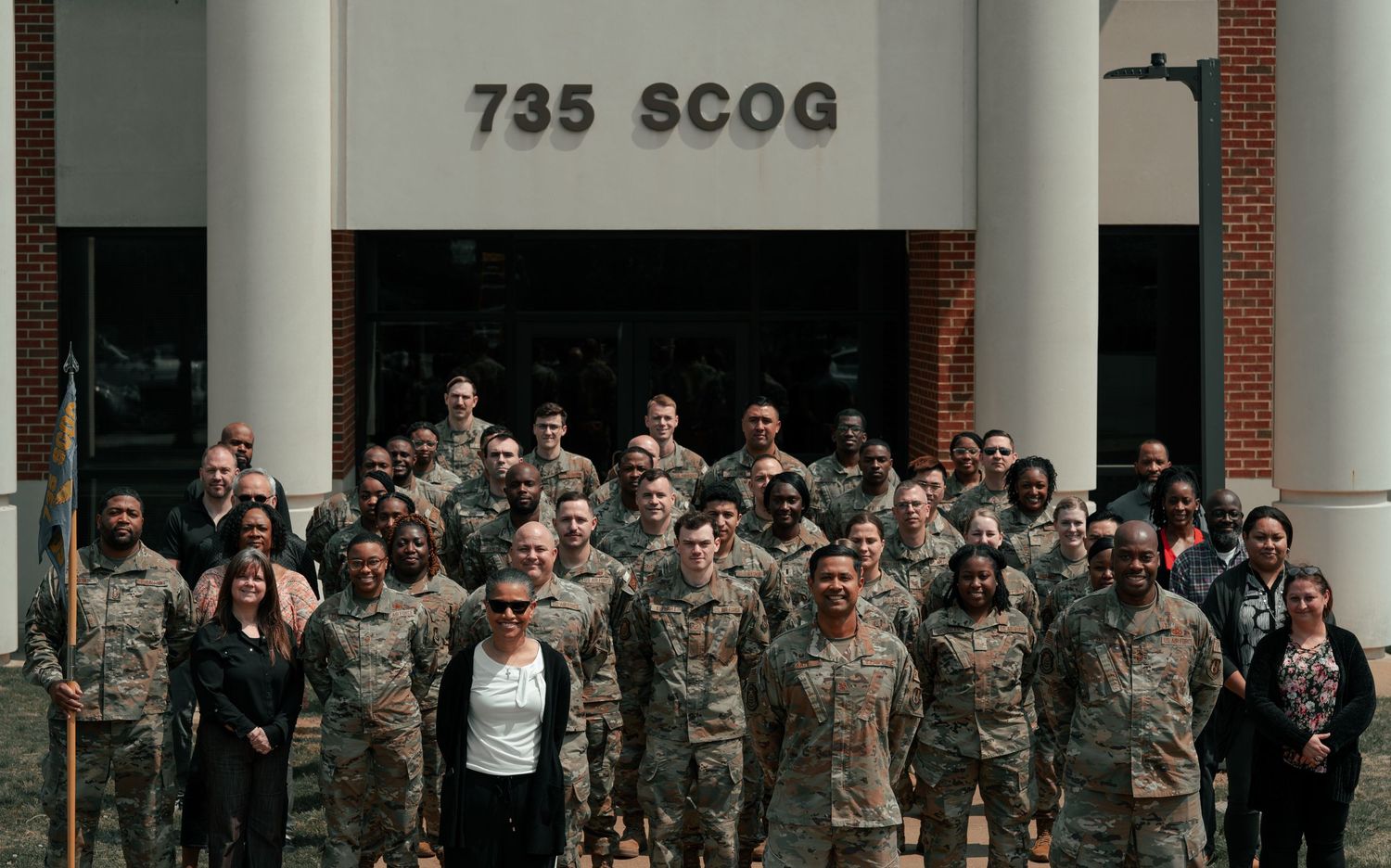 Large group of military personnel and civilians pose for group photo outside building entrance.