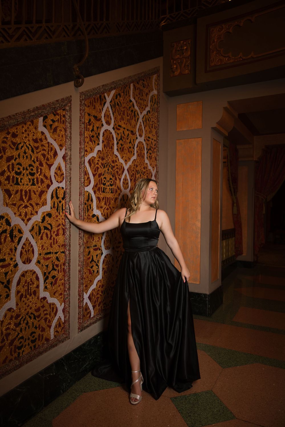 Woman in elegant black slit dress poses against ornate decorative wall panel in vintage interior setting.