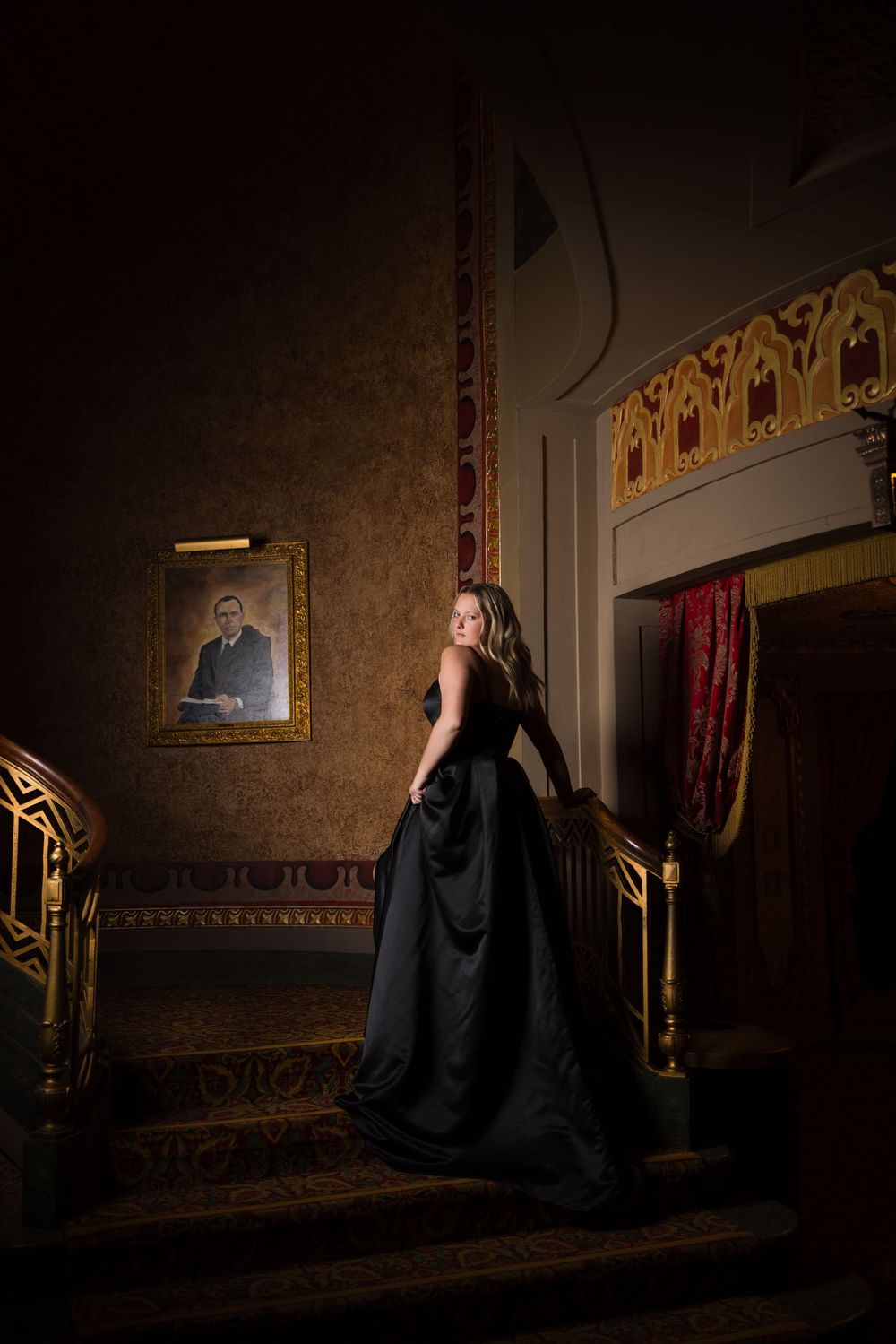 A woman in a glamorous black ballgown poses on elegant stairs in a vintage-styled room with ornate decor.