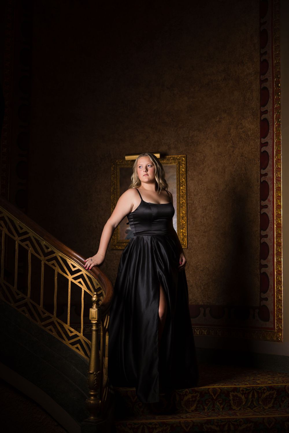 A figure in a long black formal gown stands elegantly on an ornate staircase with a vintage gold frame in the background.