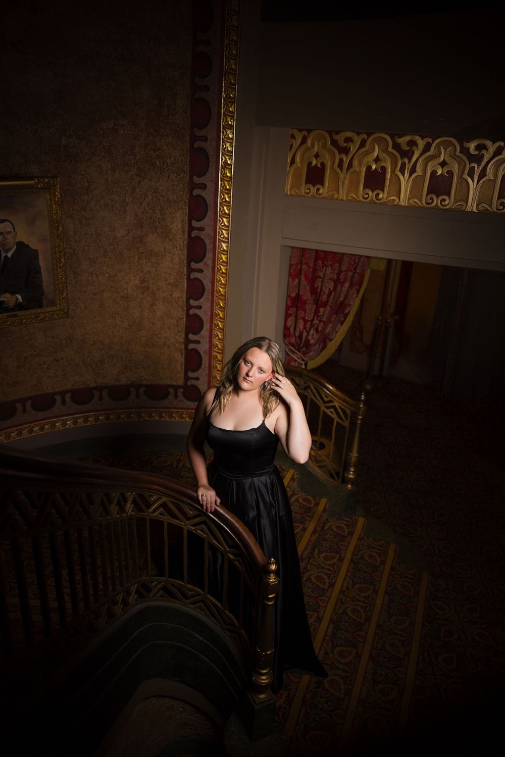 A person in an elegant black dress stands on ornate vintage stairs with decorative gold details in moody lighting.