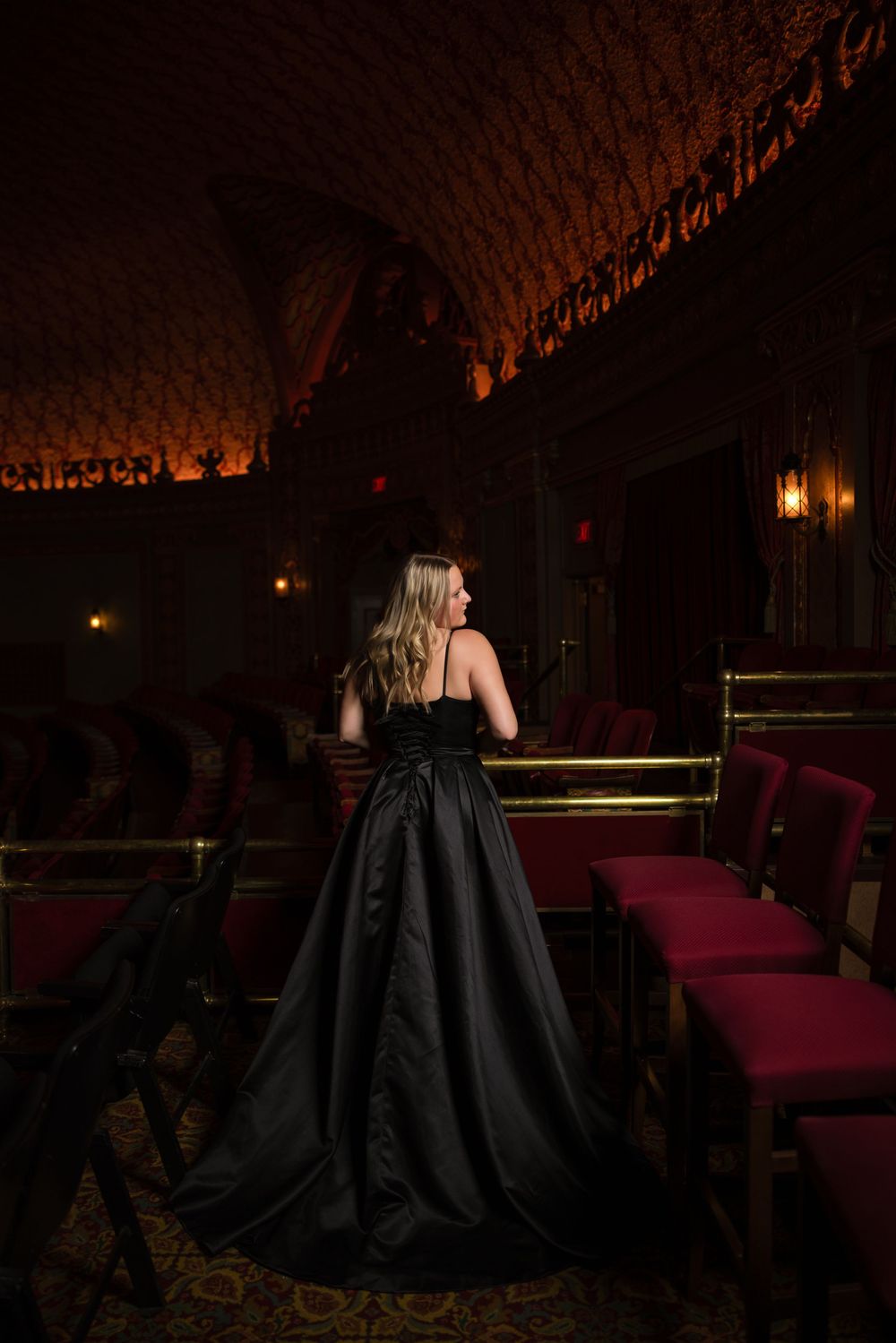 Woman in elegant black ball gown poses in dark vintage theater with ornate ceiling and dramatic lighting.