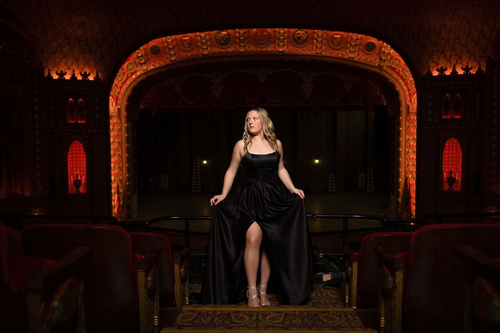 Elegant figure in a flowing black dress poses dramatically on ornate theater stage with vintage red and gold archway.