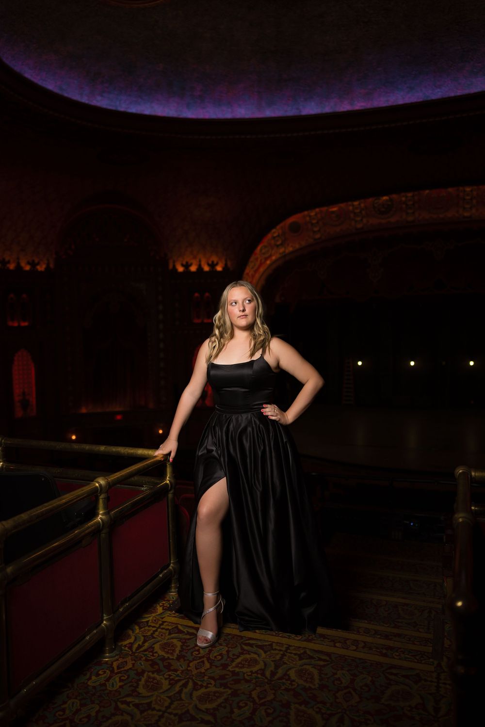 A woman in a black evening gown poses elegantly on a balcony in an ornate theater with vintage decor and lighting.