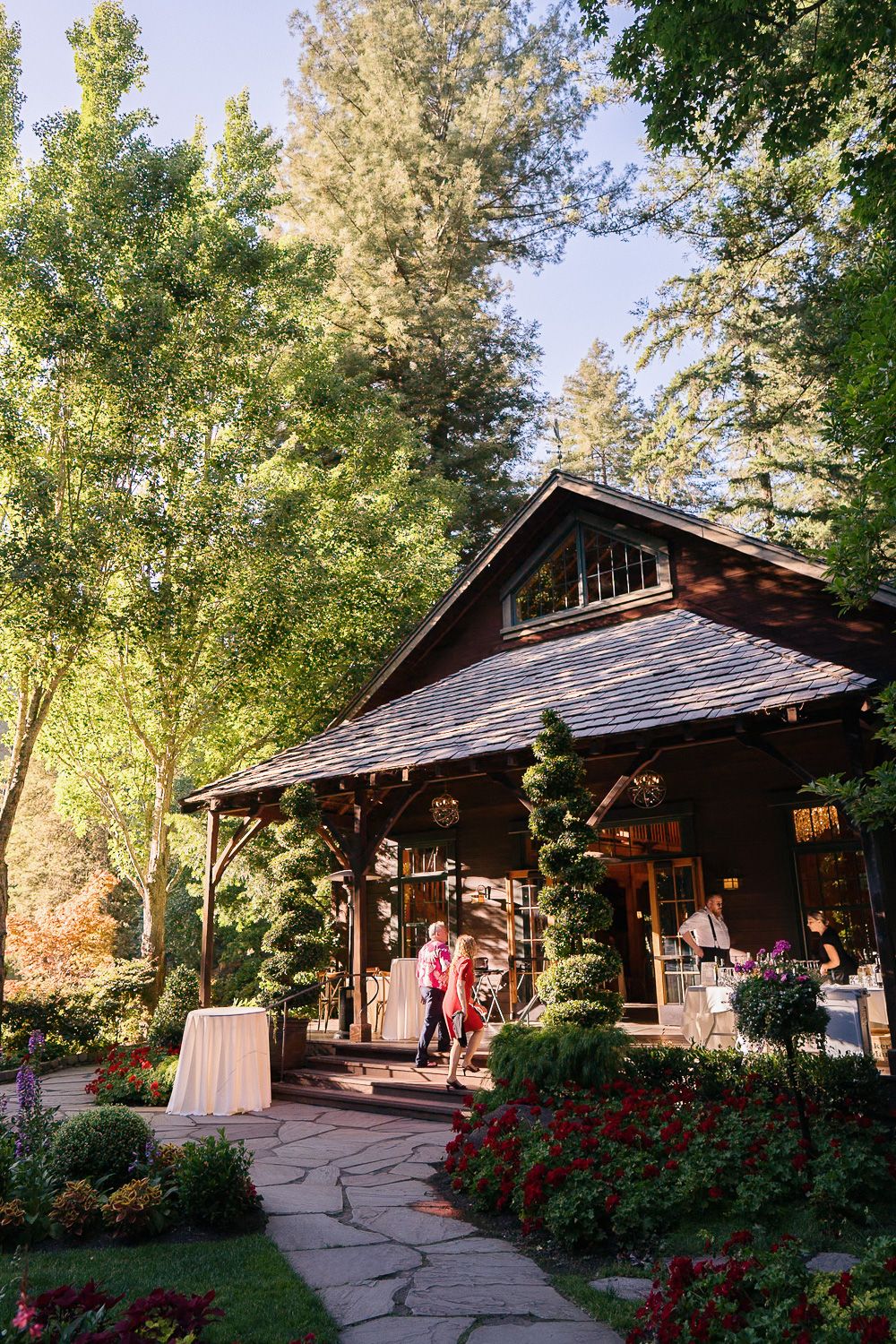 A rustic wooden cabin venue with stone pathway and red flowers surrounded by tall trees in a garden setting.