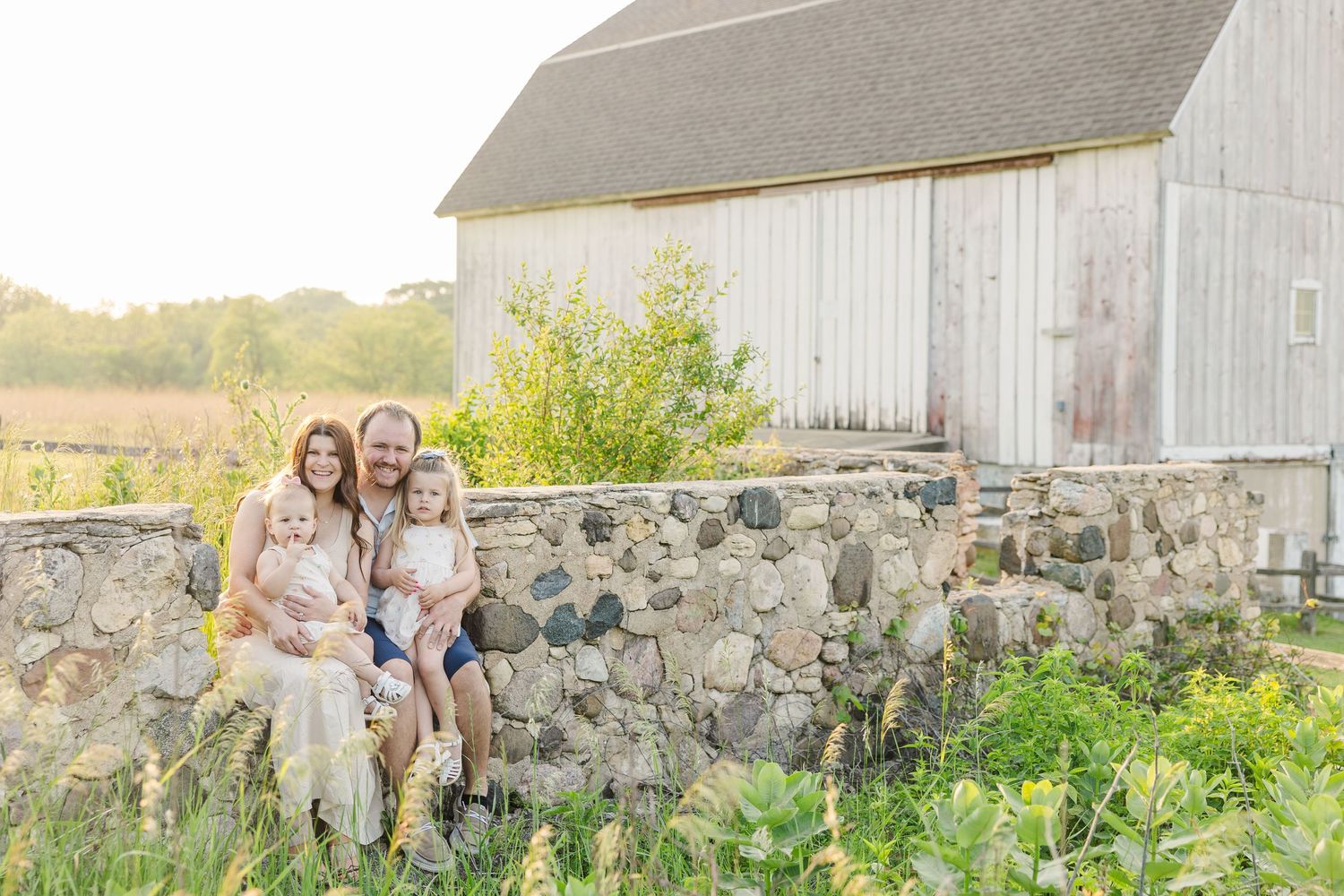 A family sits together on a rustic stone wall in front of a white barn during golden hour in nature.