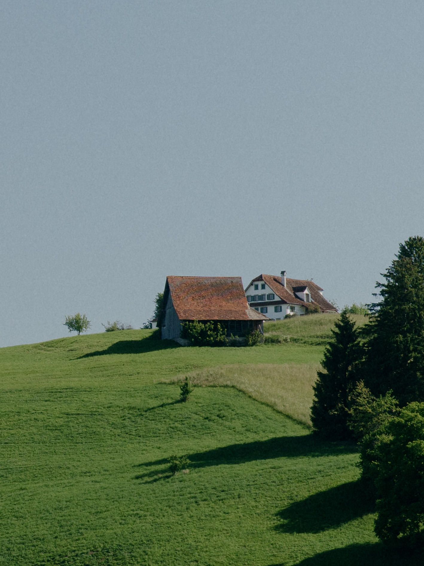 Rustic farmhouse and barn sit atop green rolling hills against a misty blue sky in a pastoral countryside scene.