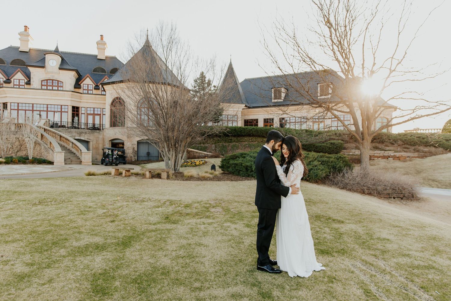 A romantic sunset wedding photo of a couple embracing on a manicured lawn in front of an elegant mansion estate.