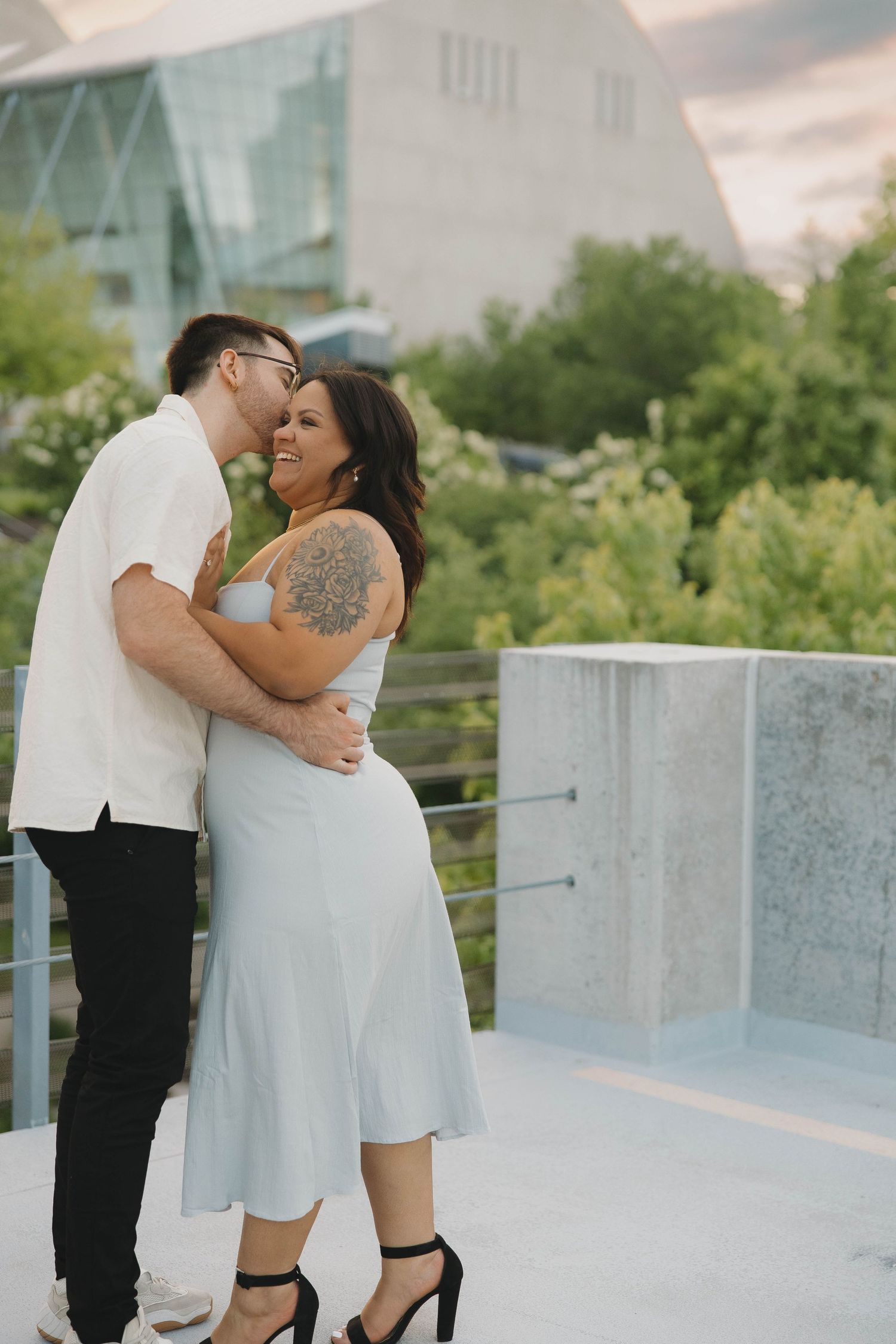 A couple embraces intimately on an outdoor terrace with greenery and modern architecture in the background.
