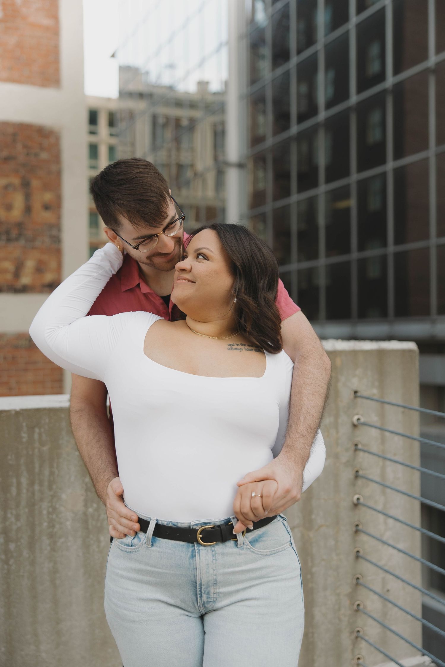 A romantic couple embraces against a brick and glass building exterior in an urban setting.