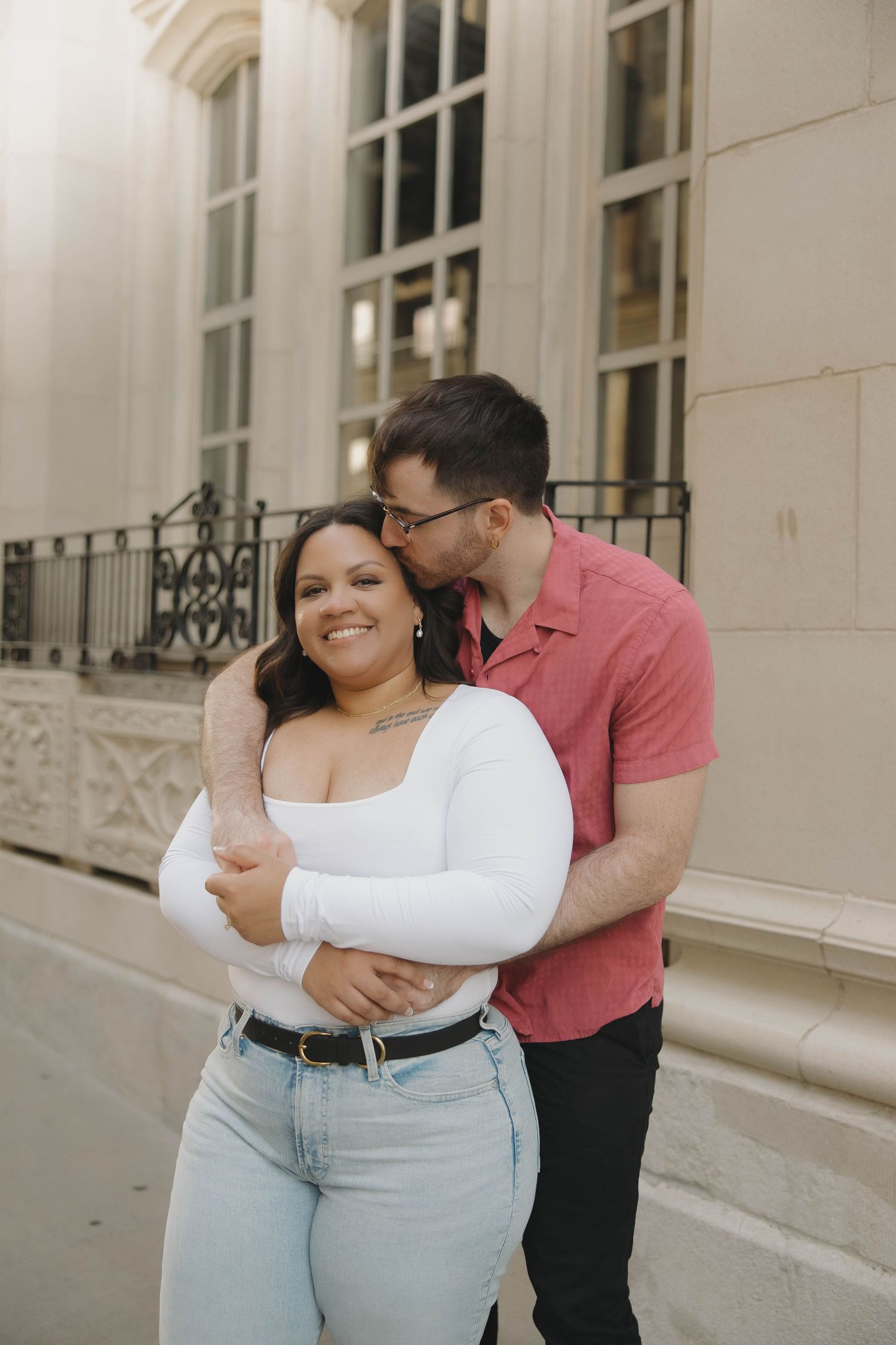 A romantic couple embraces and shares affectionate moments together against a light colored building exterior.
