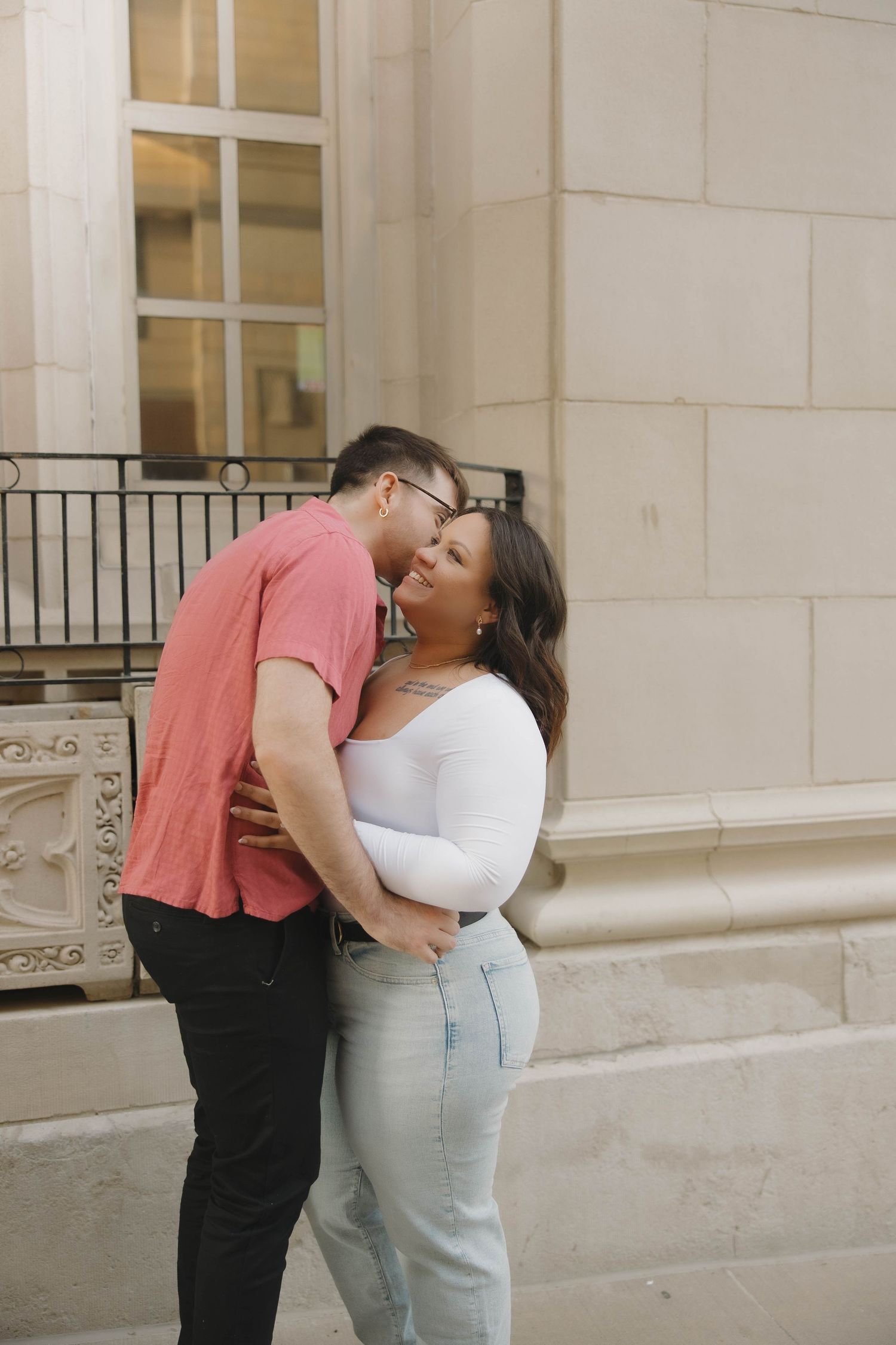 A loving couple in casual attire sharing an intimate moment against a white stone building exterior.
