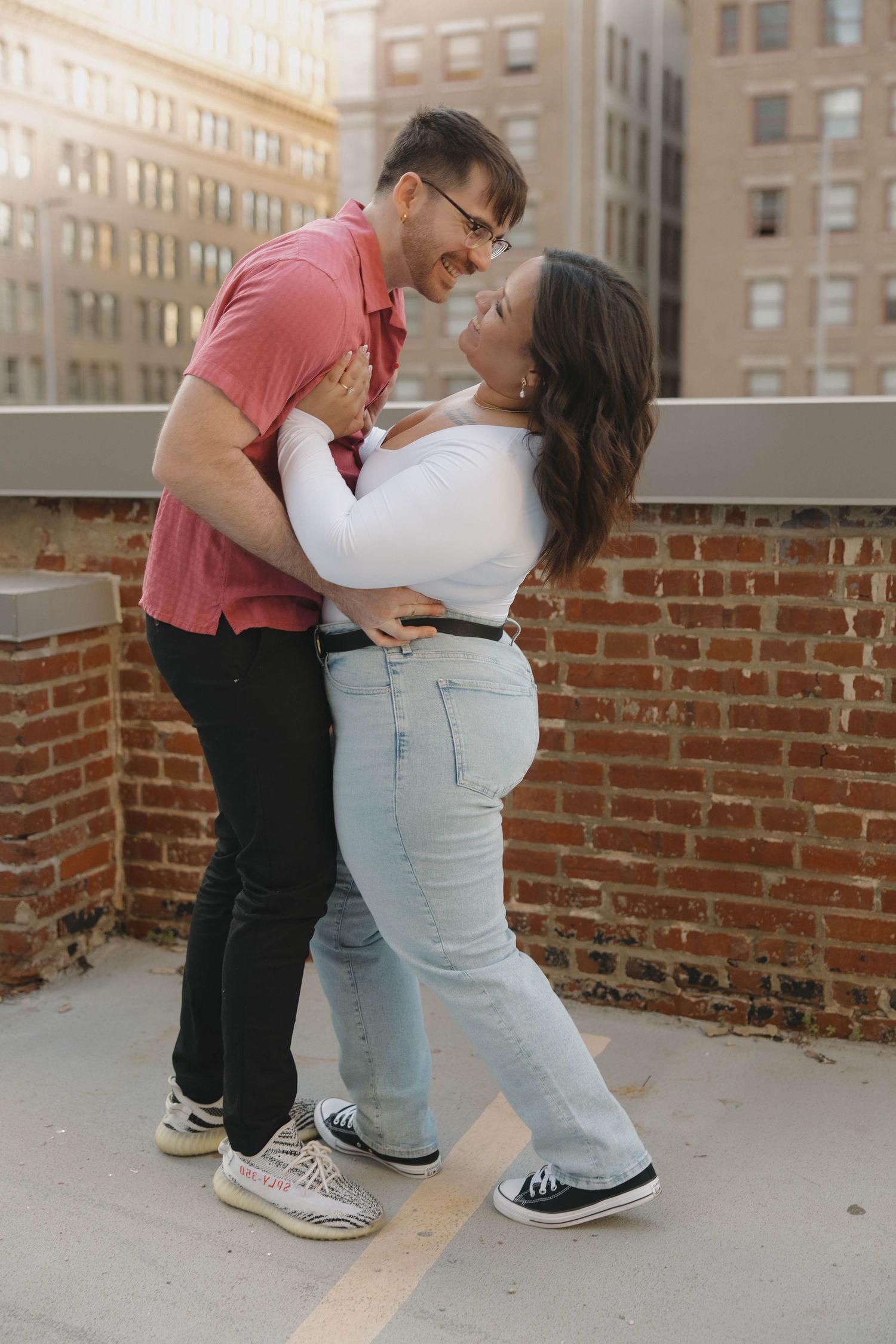 A romantic couple embraces and laughs together on a rooftop patio with brick walls and city buildings in the background.