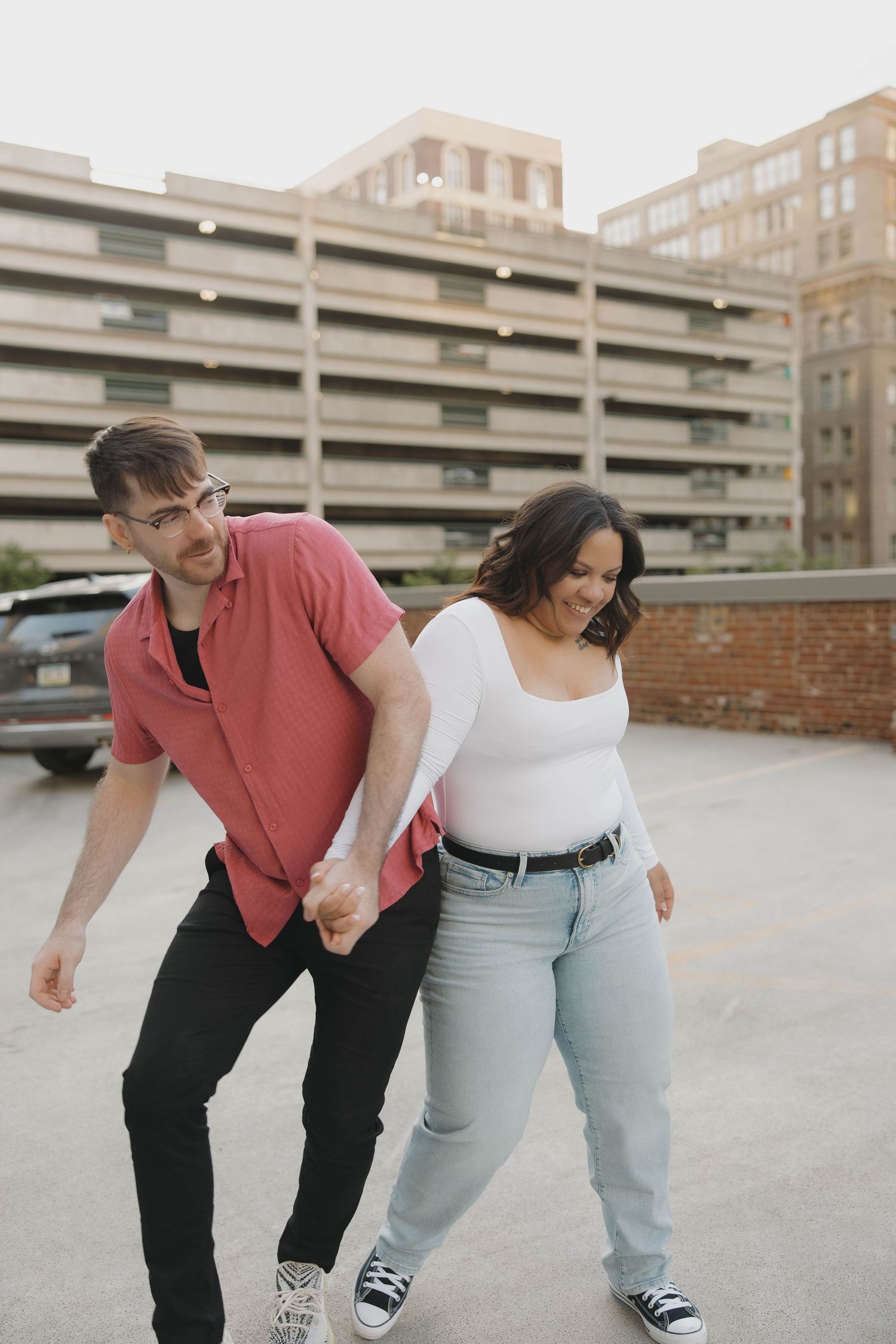 Couple holding hands and walking playfully in urban parking lot wearing casual outfits.