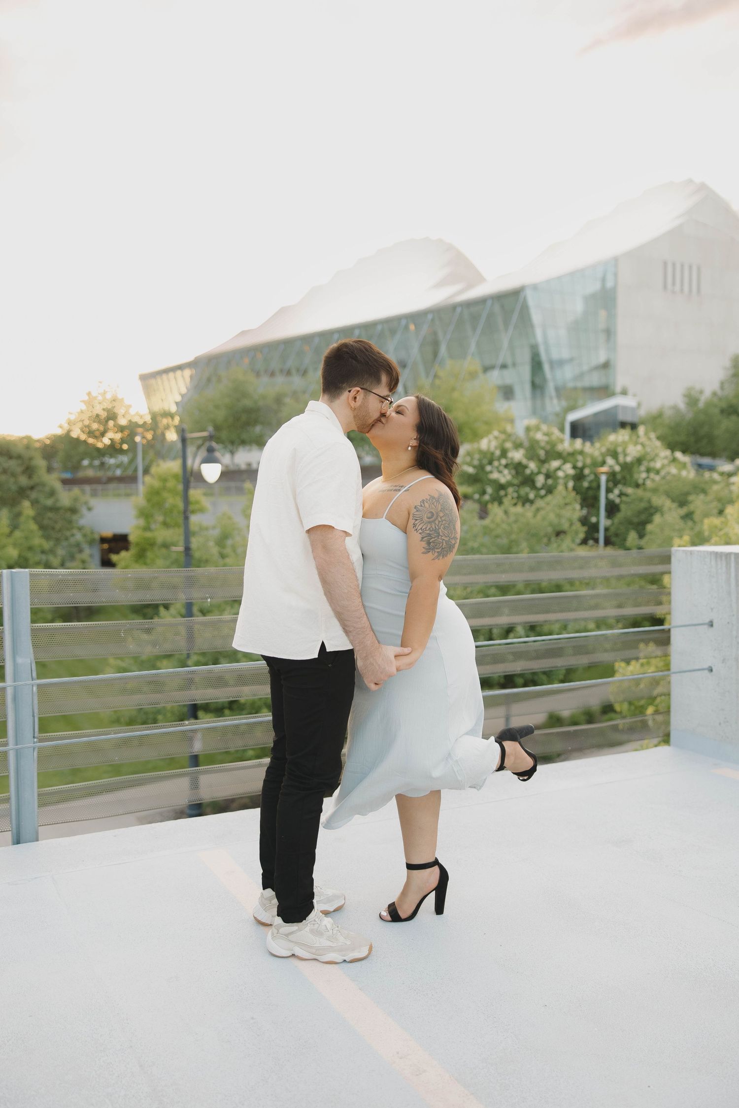 A romantic couple embraces and kisses while standing on a rooftop deck with a modern building in the background.