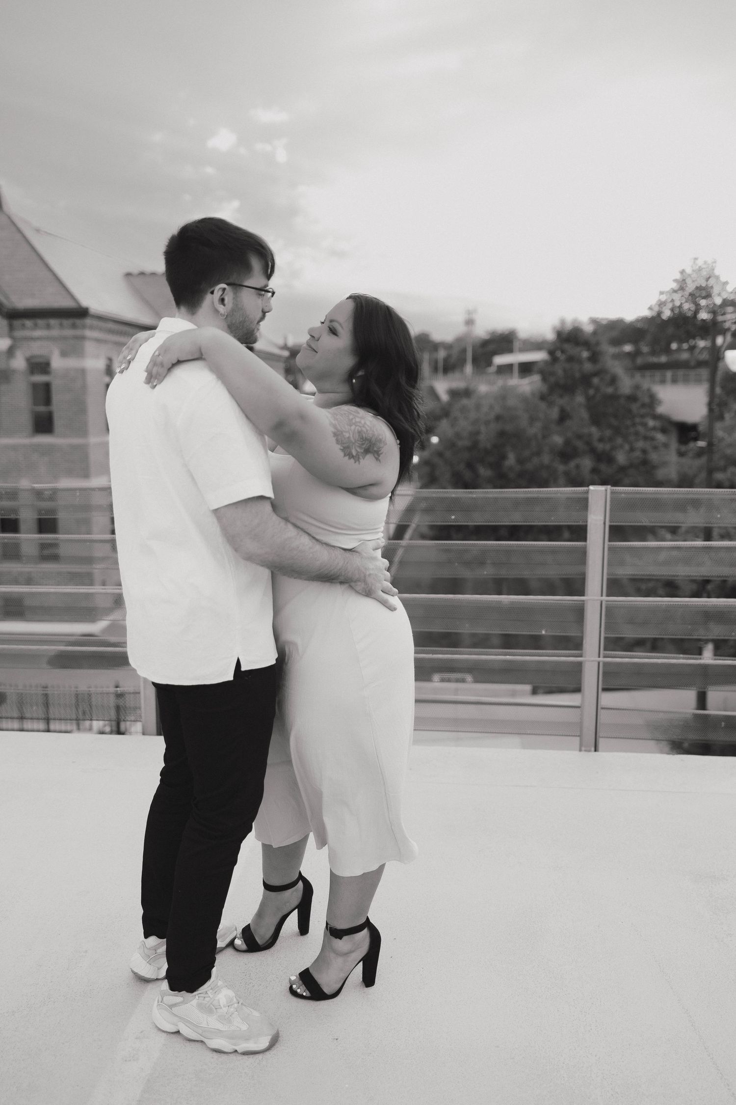 A romantic couple embraces in black and white on a rooftop balcony with urban scenery in the background.