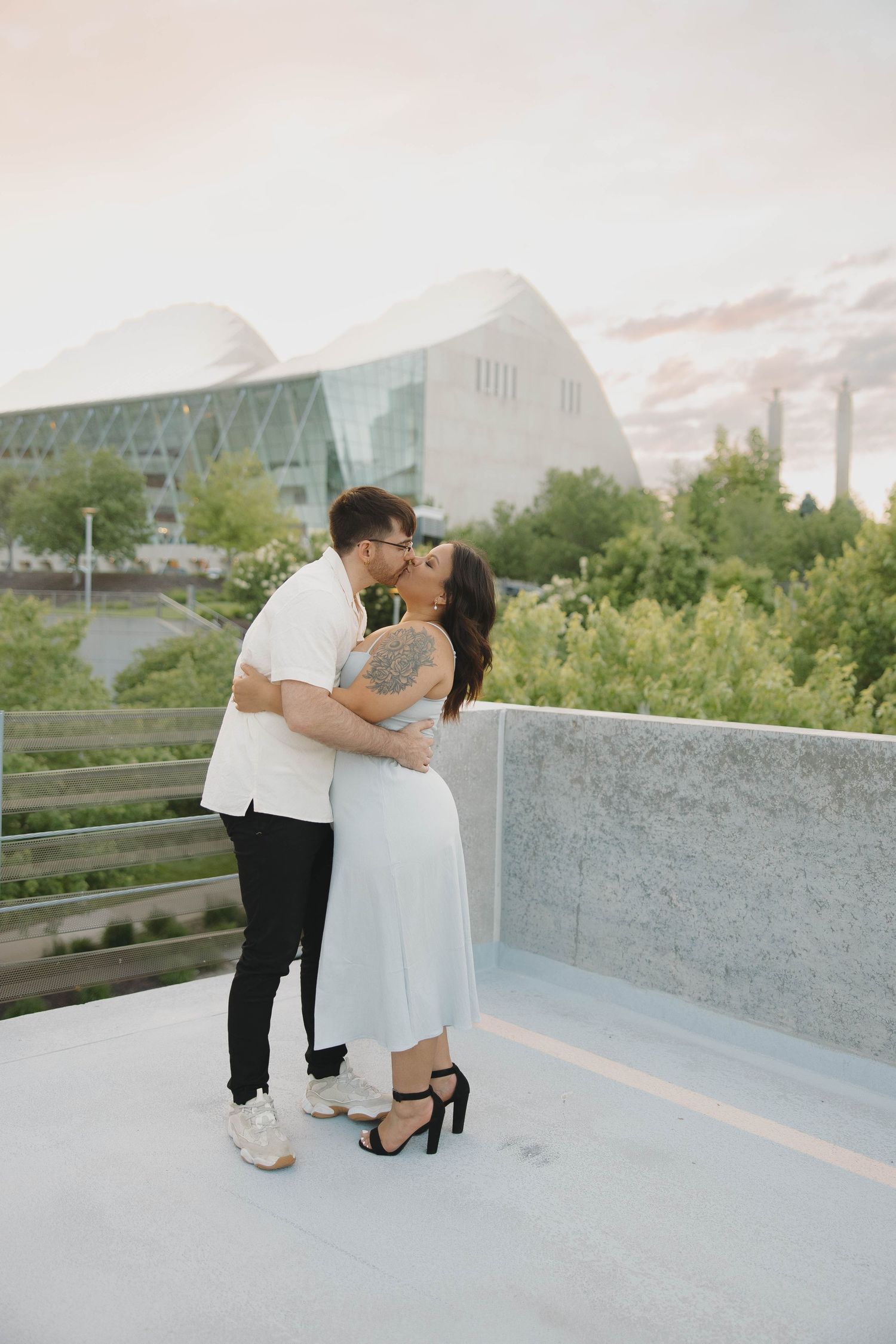 A couple shares an intimate moment on an urban rooftop with modern architectural buildings in the background.