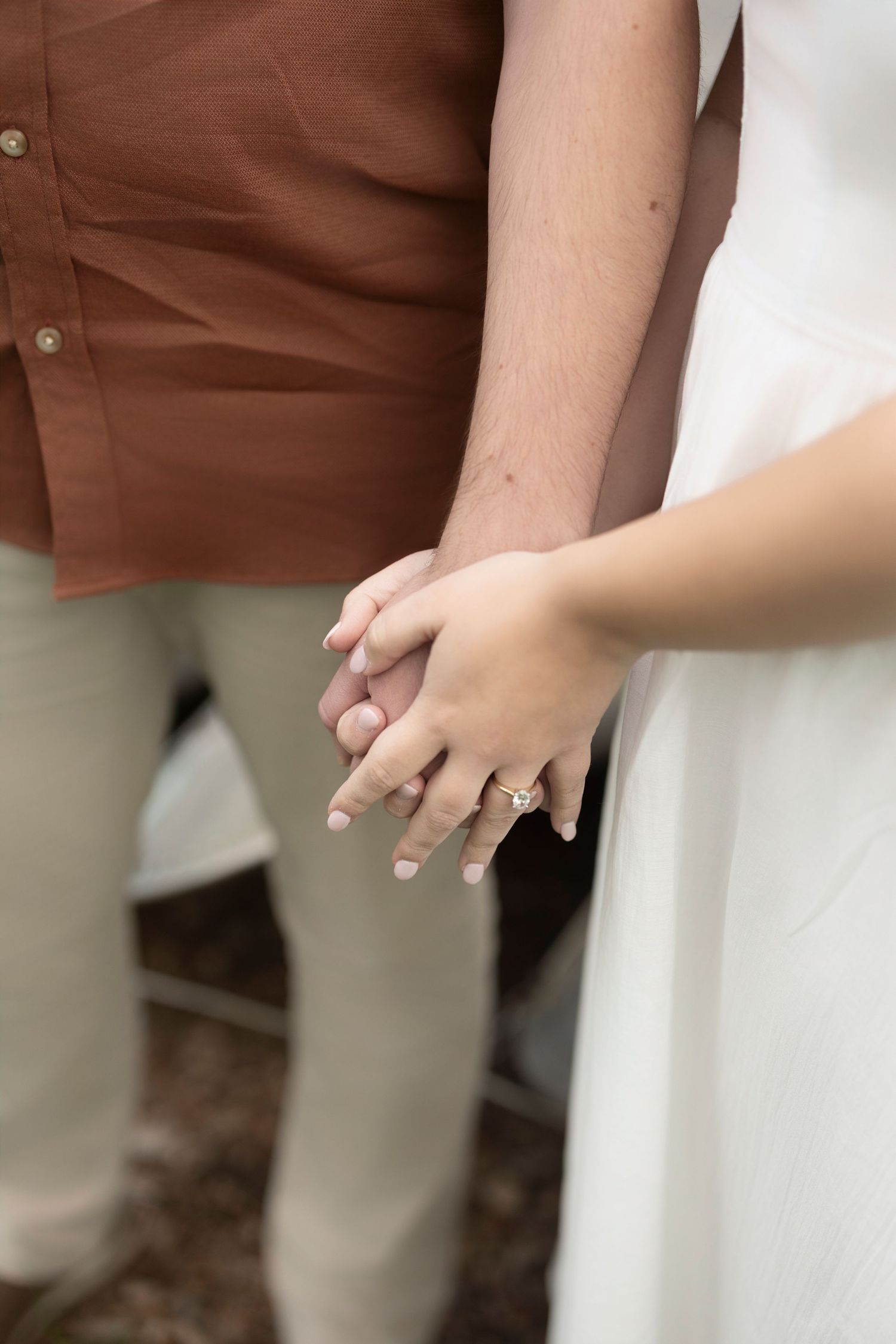 Close up of two clasped hands with an engagement ring visible against brown and white clothing.