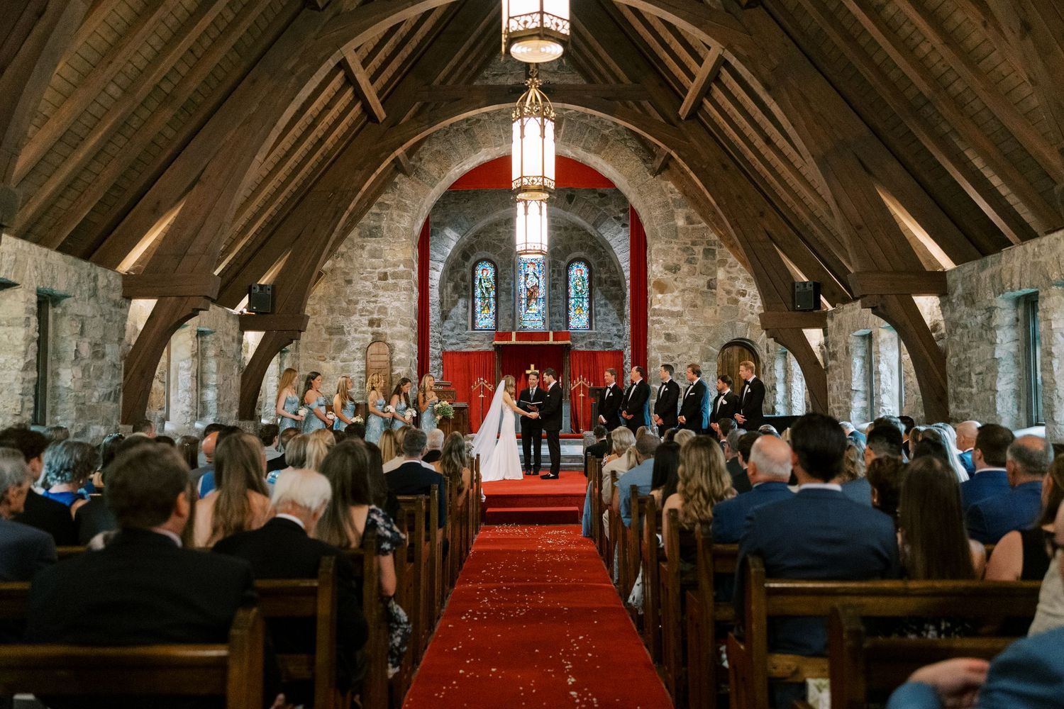 Wedding ceremony inside rustic stone chapel at Silver Bay YMCA with vaulted wooden ceiling, red carpet aisle and stained glass windows.