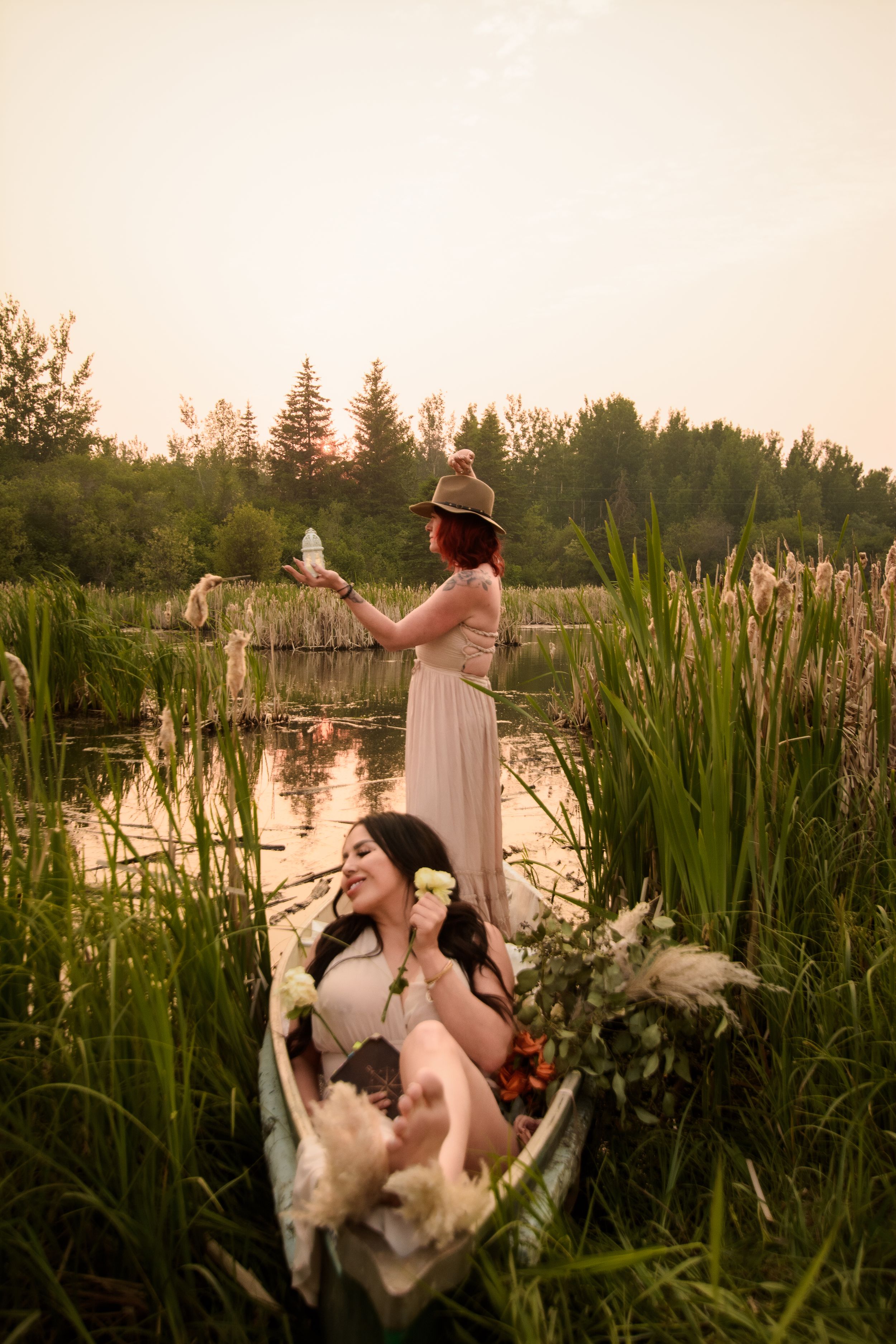 Scenic sunset photo of two people in a wooden boat among tall marsh reeds and water lilies at golden hour.
