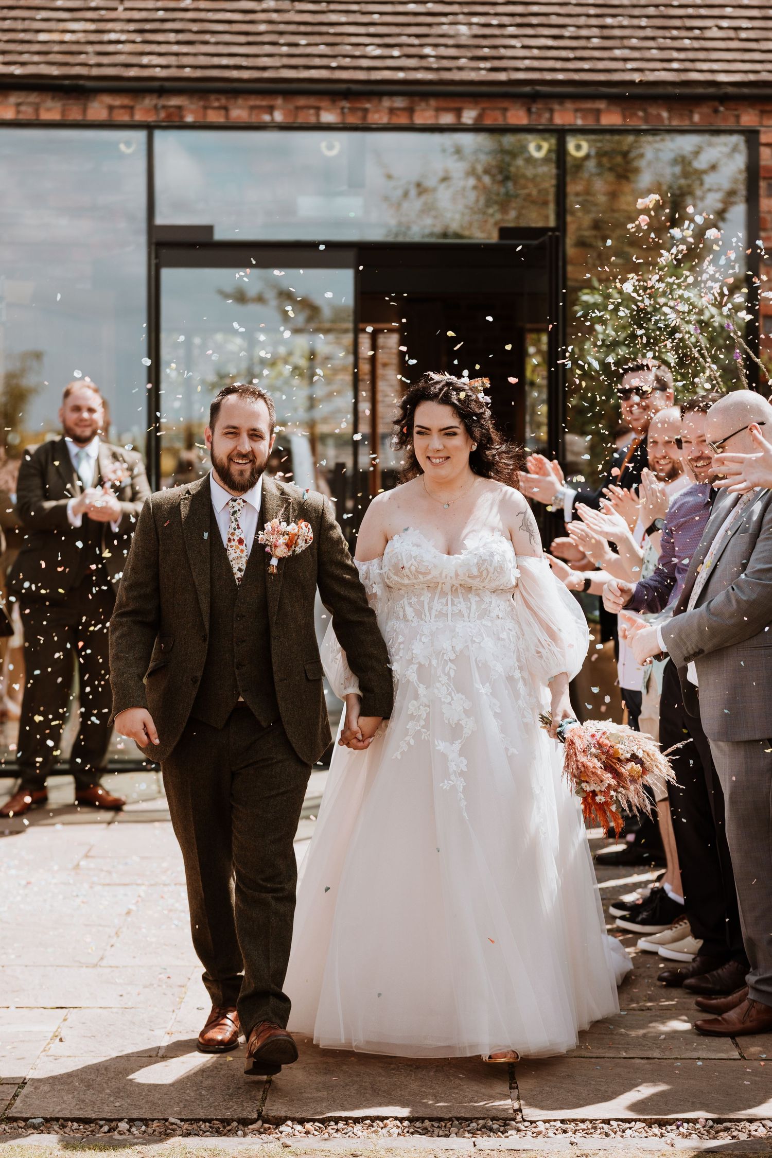 A couple exits their wedding venue while guests shower them with confetti on a sunny autumn day at Curradine Barns
