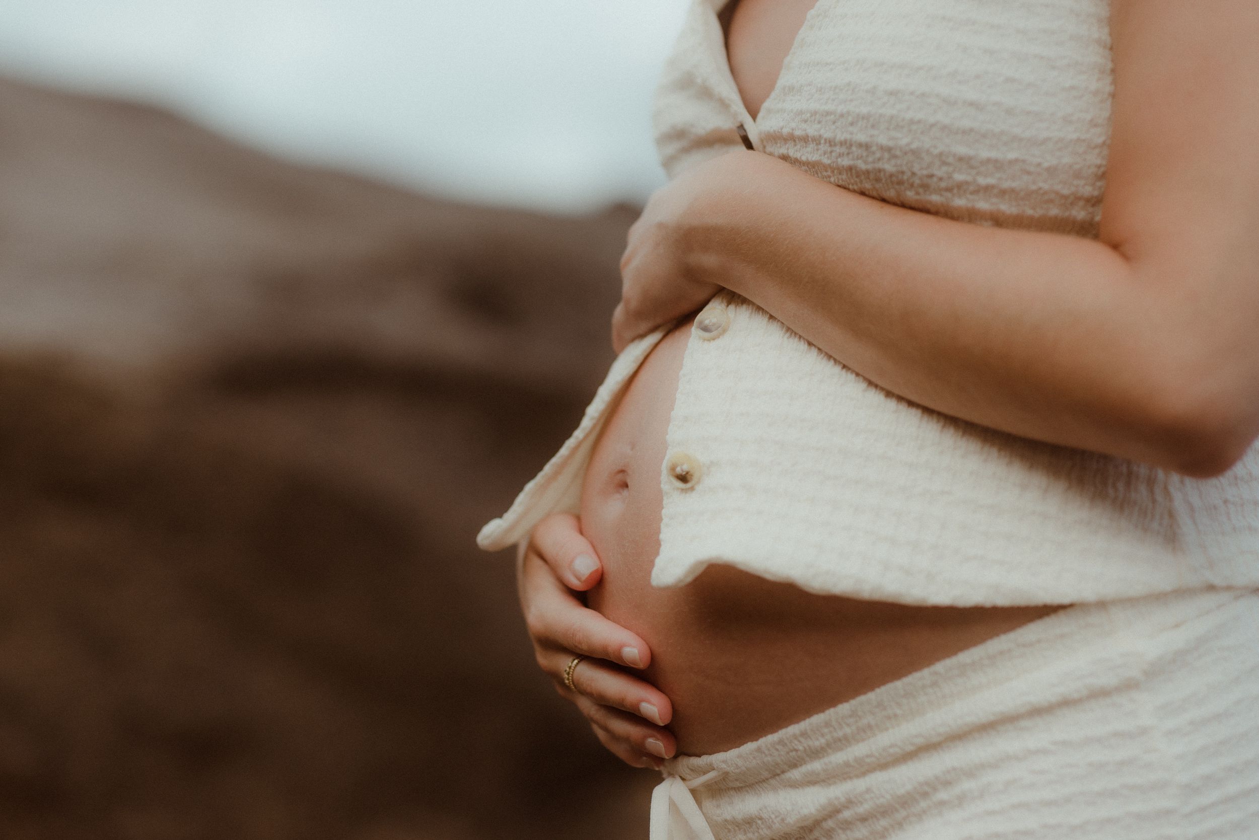 A pregnant woman in a white dress tenderly cradles their baby bump against a blurred background at sunset in El Médano, Tenerife.