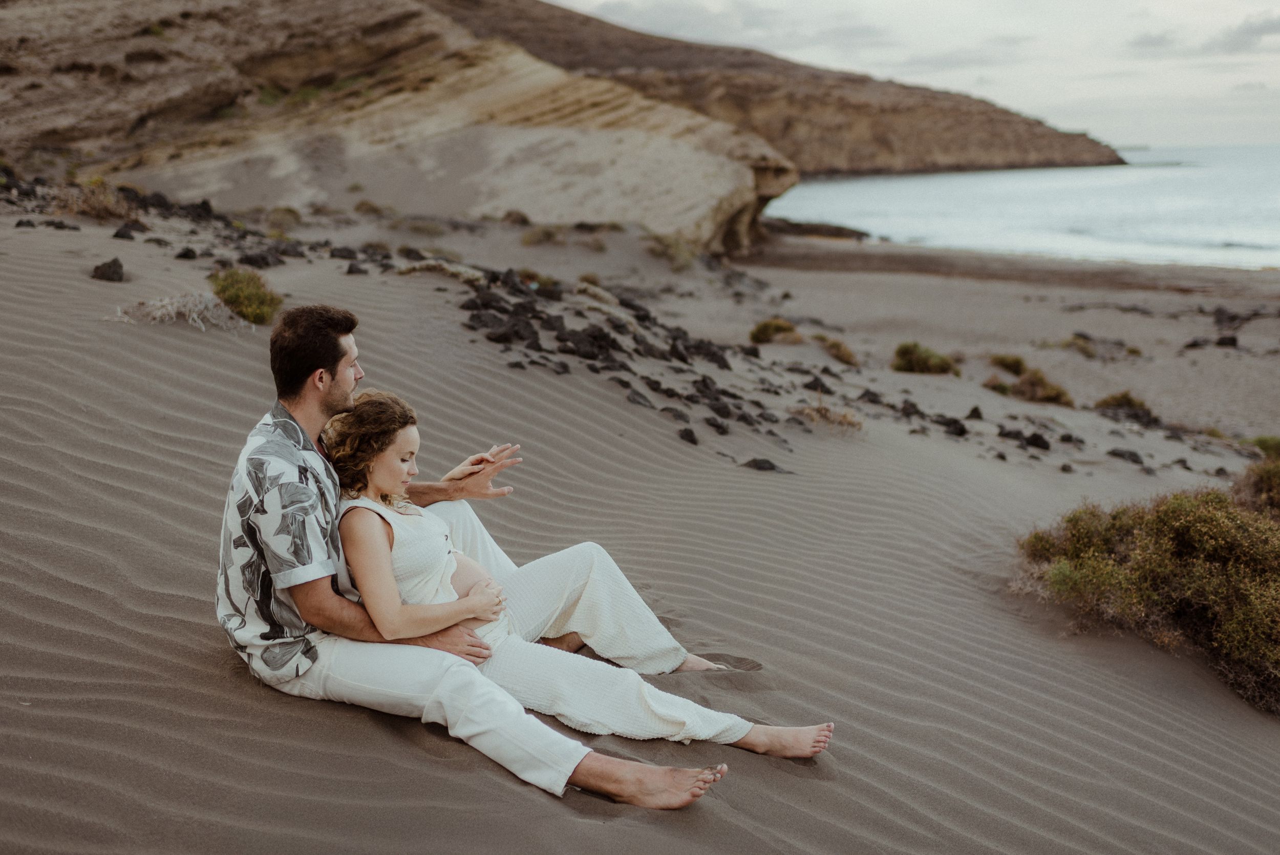 Couple in elegant attire relaxes together on a scenic beach with dramatic cliffs and coastline in the background in El Médano, Tenerife.