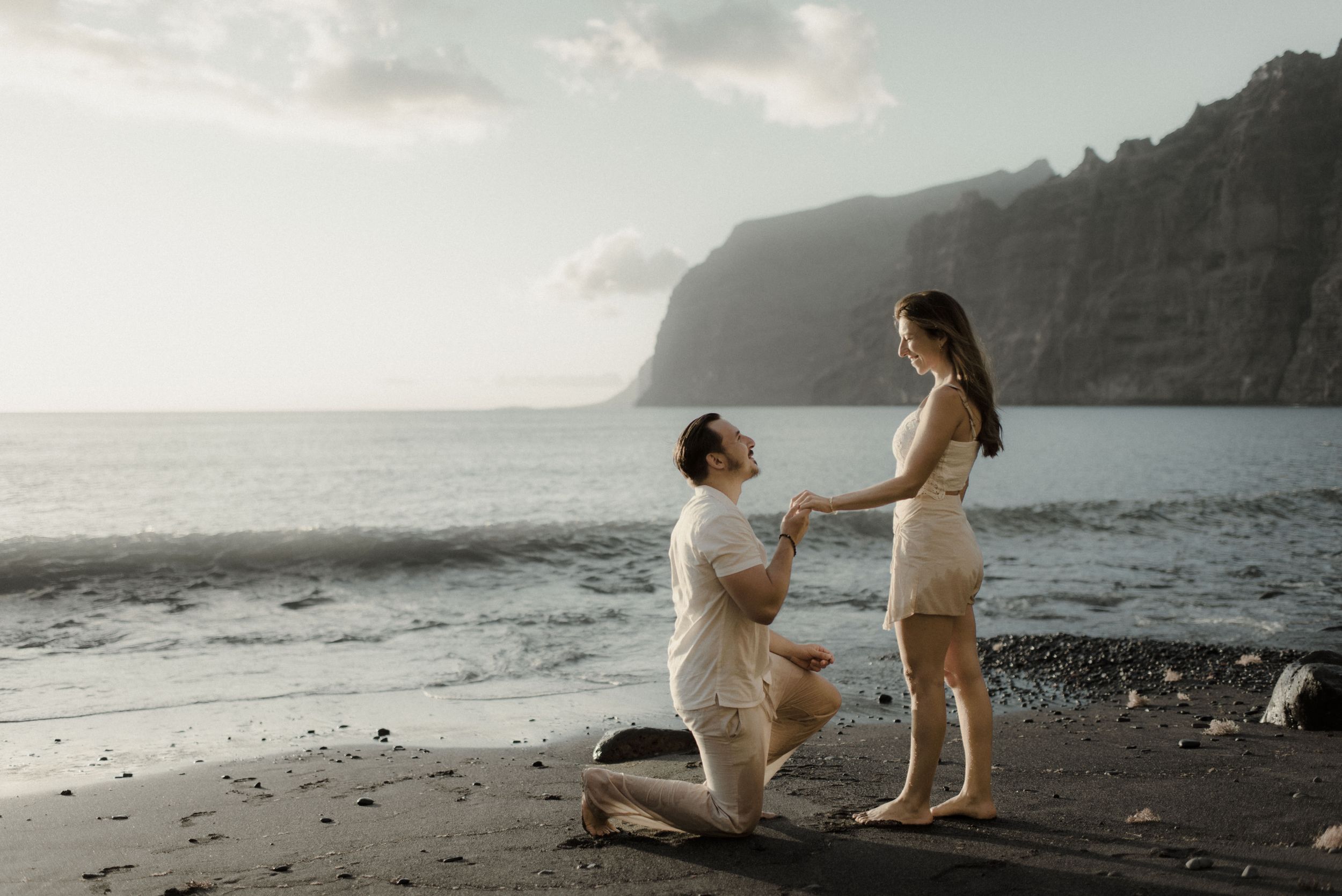 A romantic proposal scene with figures silhouetted against dramatic coastal cliffs and ocean at sunset on a black sand beach.