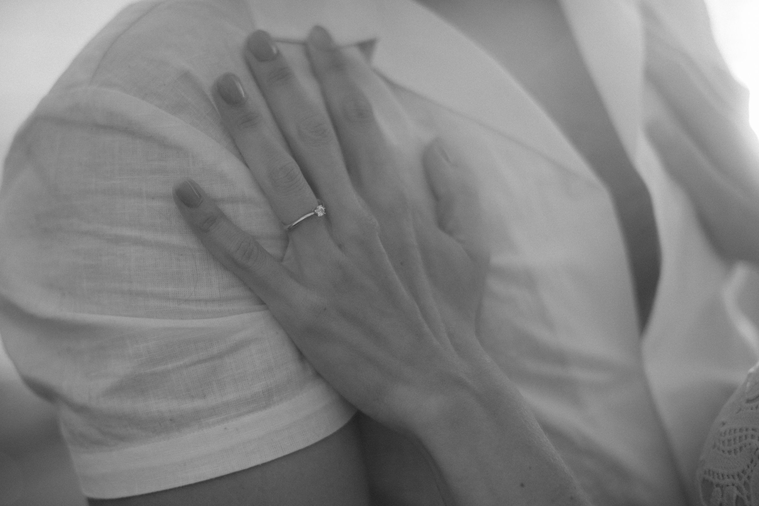 A close up black and white photograph shows a delicate hand with a wedding ring resting gently on white fabric.