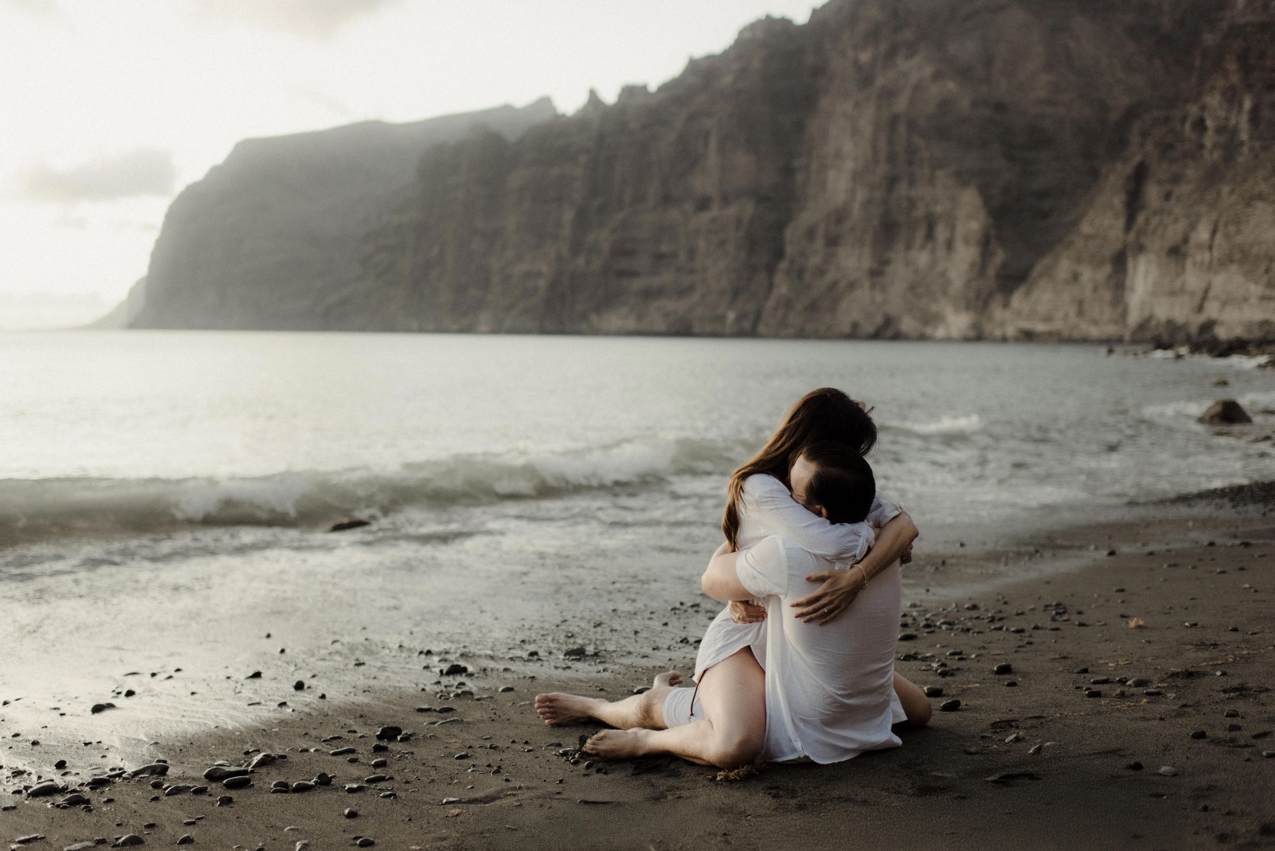 A couple embraces on a misty beach with dramatic cliffs and rocky coastline in the background.