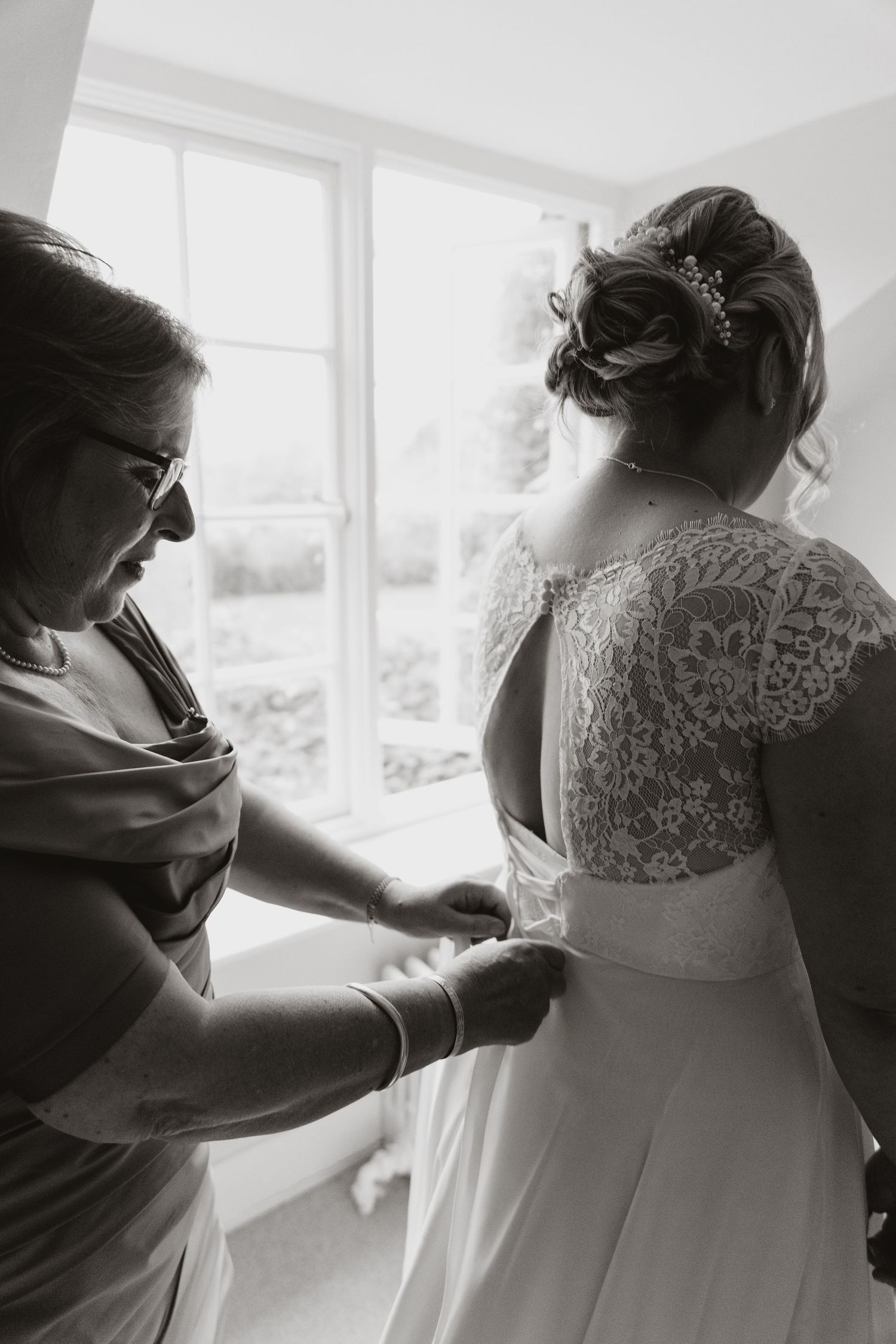 A black and white photograph of a wedding dress being adjusted near a bright window.