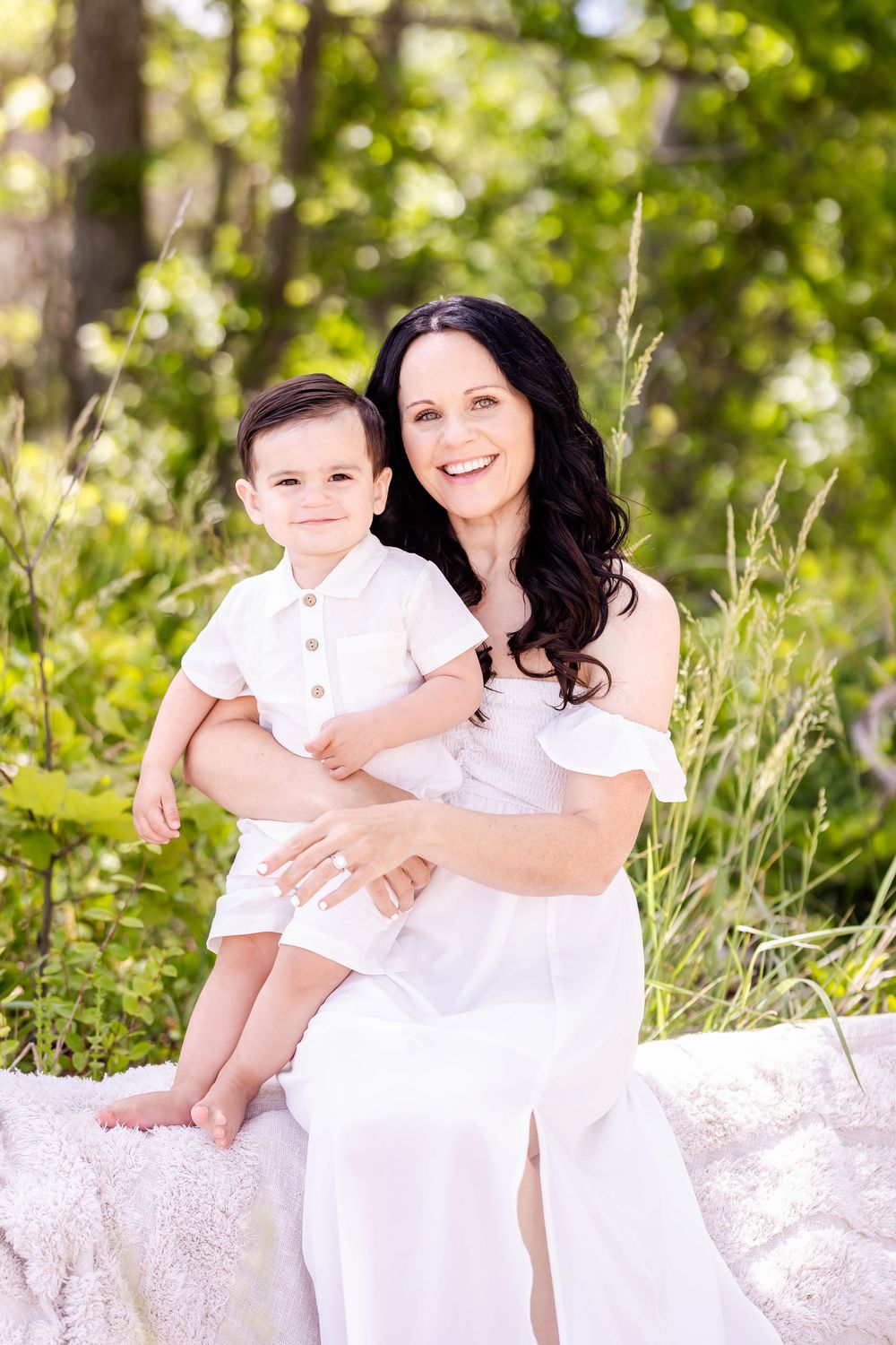 Portrait of a mother and child sitting together outdoors in white clothing on a sunny day with green foliage background.