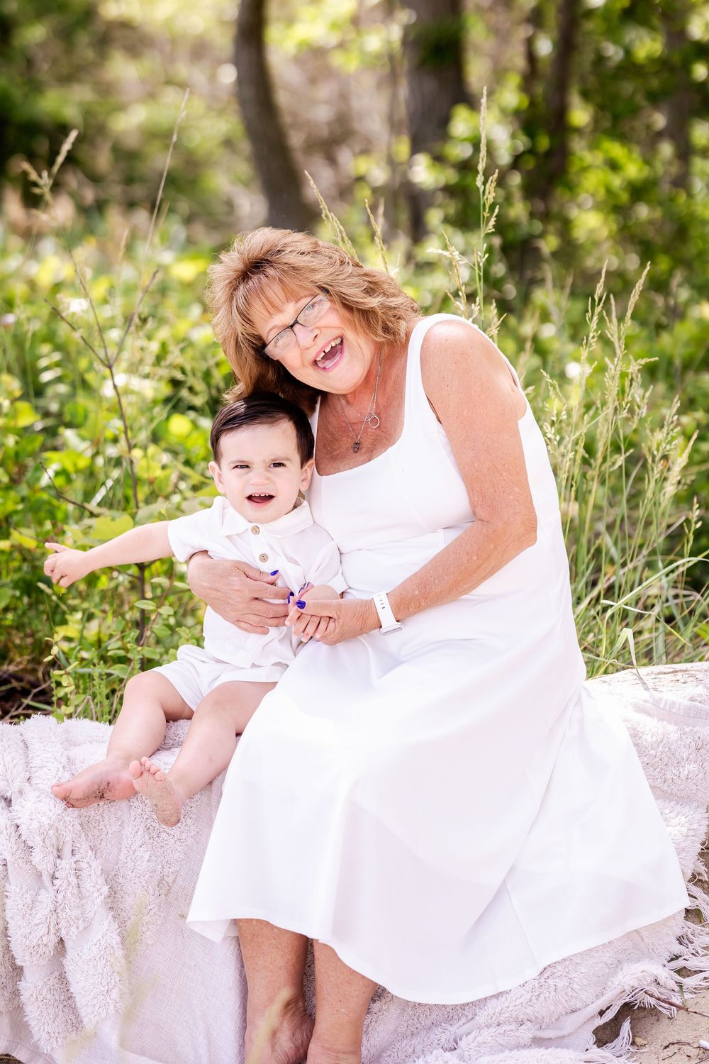 Two people wearing white dresses share joyful moments together on an outdoor bench surrounded by greenery.