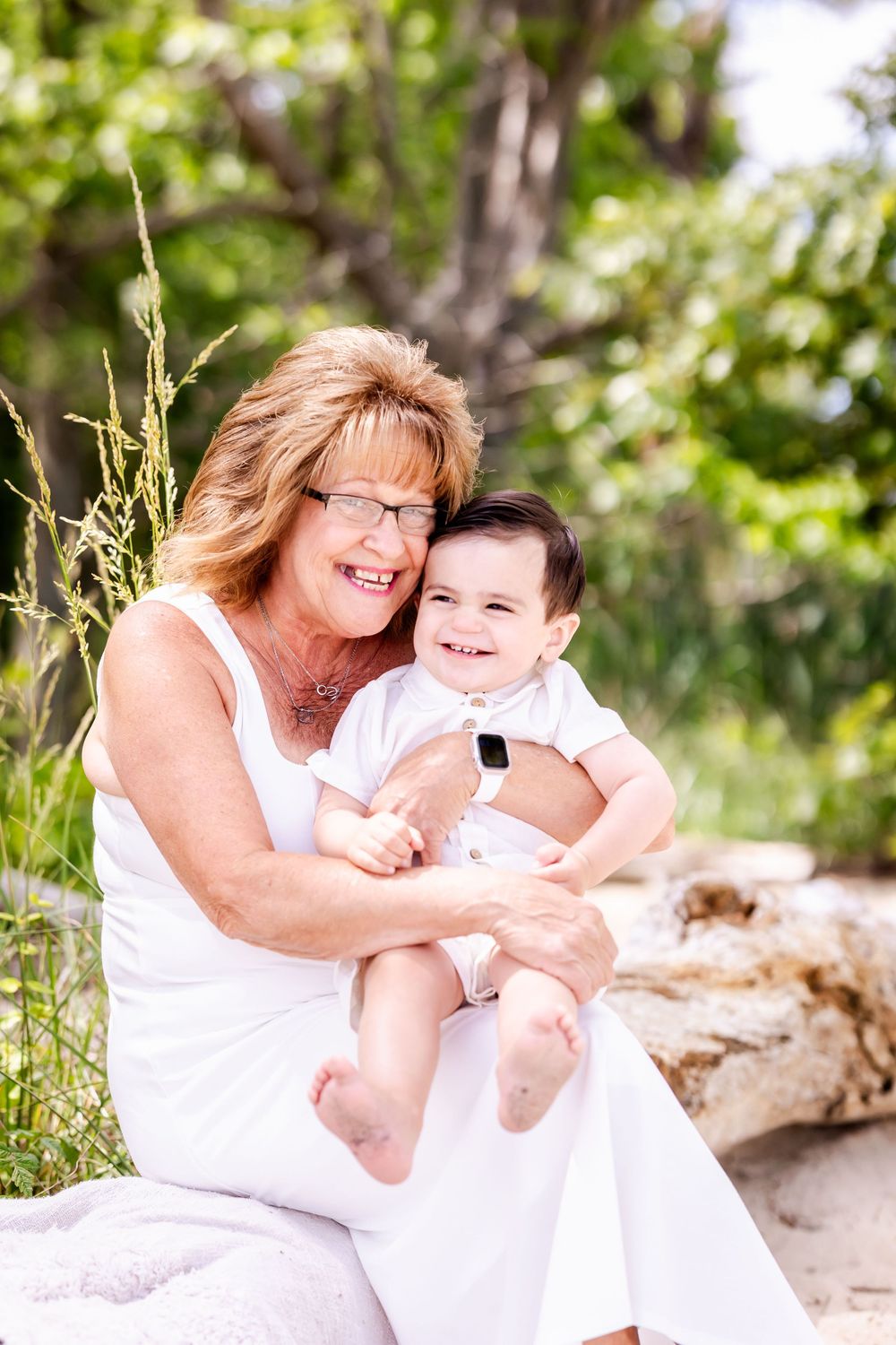A woman in a white dress sits outdoors holding a happy baby against a nature background.
