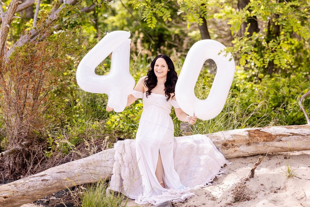 A woman in a long white dress poses outdoors with large number 40 balloons against a natural backdrop.