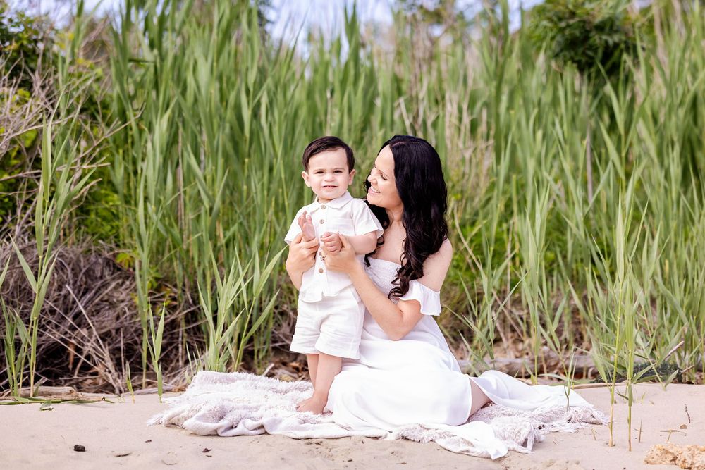 A tender moment shared between two figures sitting on a blanket at the beach amid tall beach grass on a sunny day.