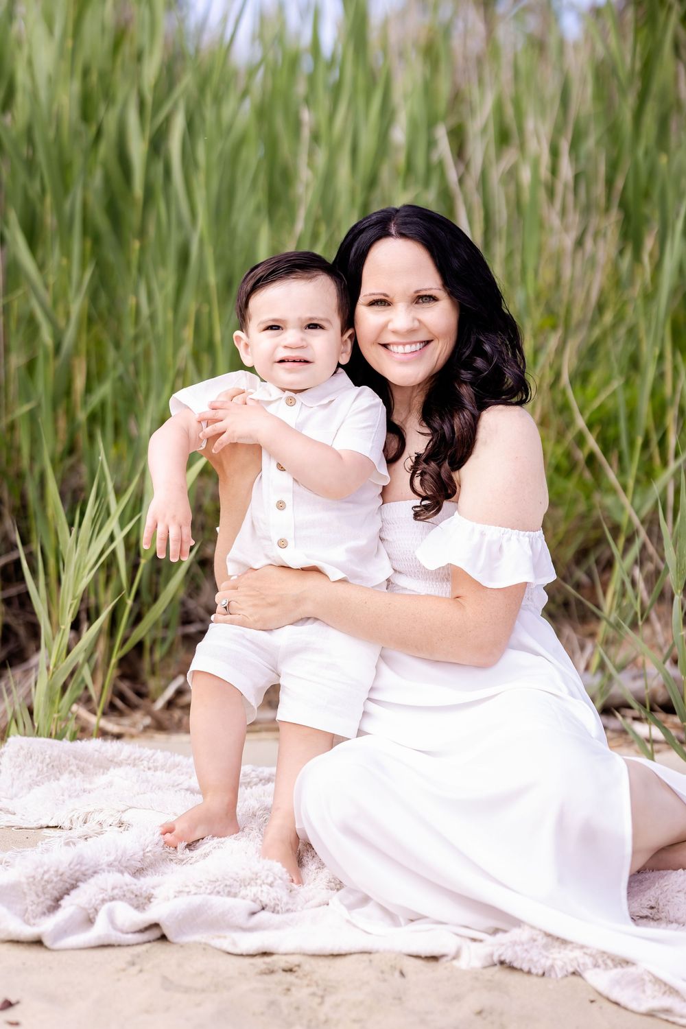 Woman in white dress sits with toddler on blanket at beach surrounded by tall grass.