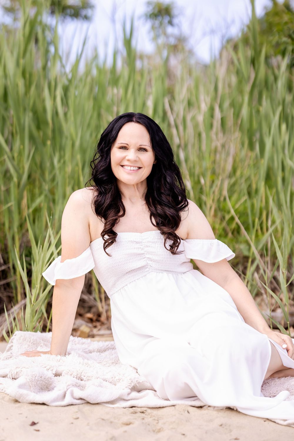 A woman in a white off-shoulder dress sits gracefully on a beach surrounded by tall grass on a sunny day.