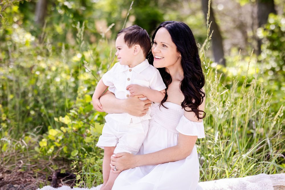 Mother in white dress holding toddler in summery outdoor setting with wildflowers and greenery in background.