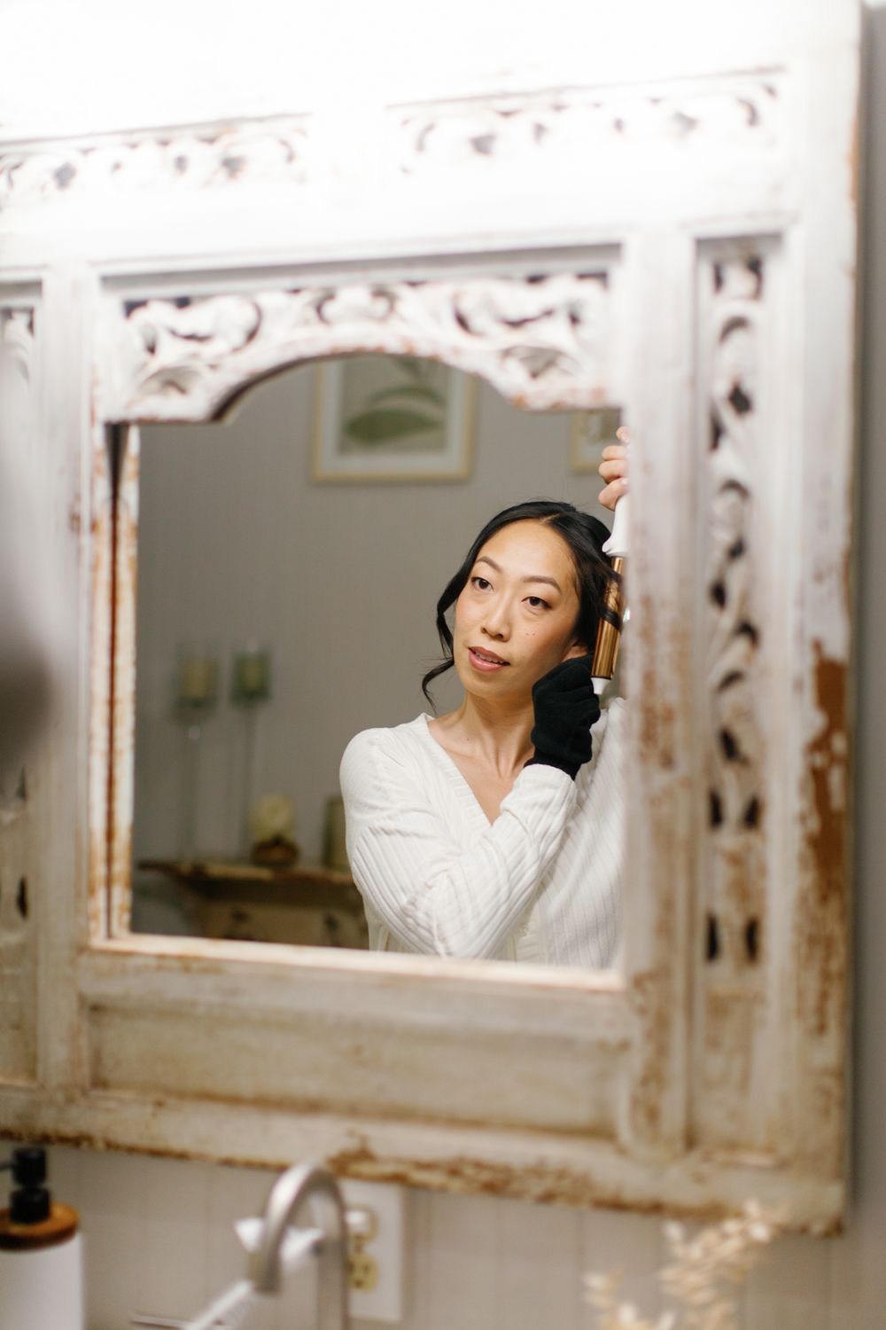 A woman in a white sweater gazes into a decorative vintage mirror with ornate floral carvings.
