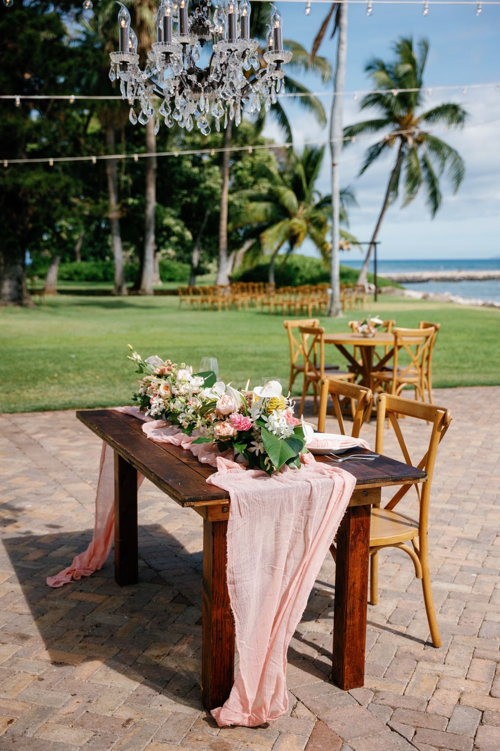 Elegant beachside table setting with pink runner and floral centerpiece under crystal chandelier by tropical palm trees.