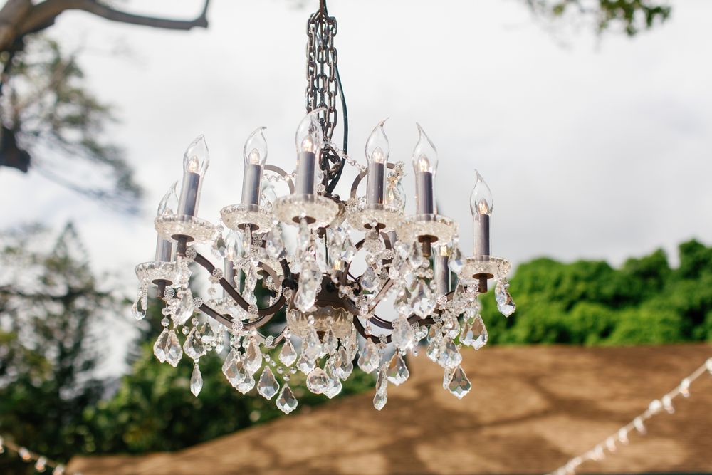 Elegant crystal chandelier with white candle-style lights and ornate glass droplets hanging outdoors against green foliage.