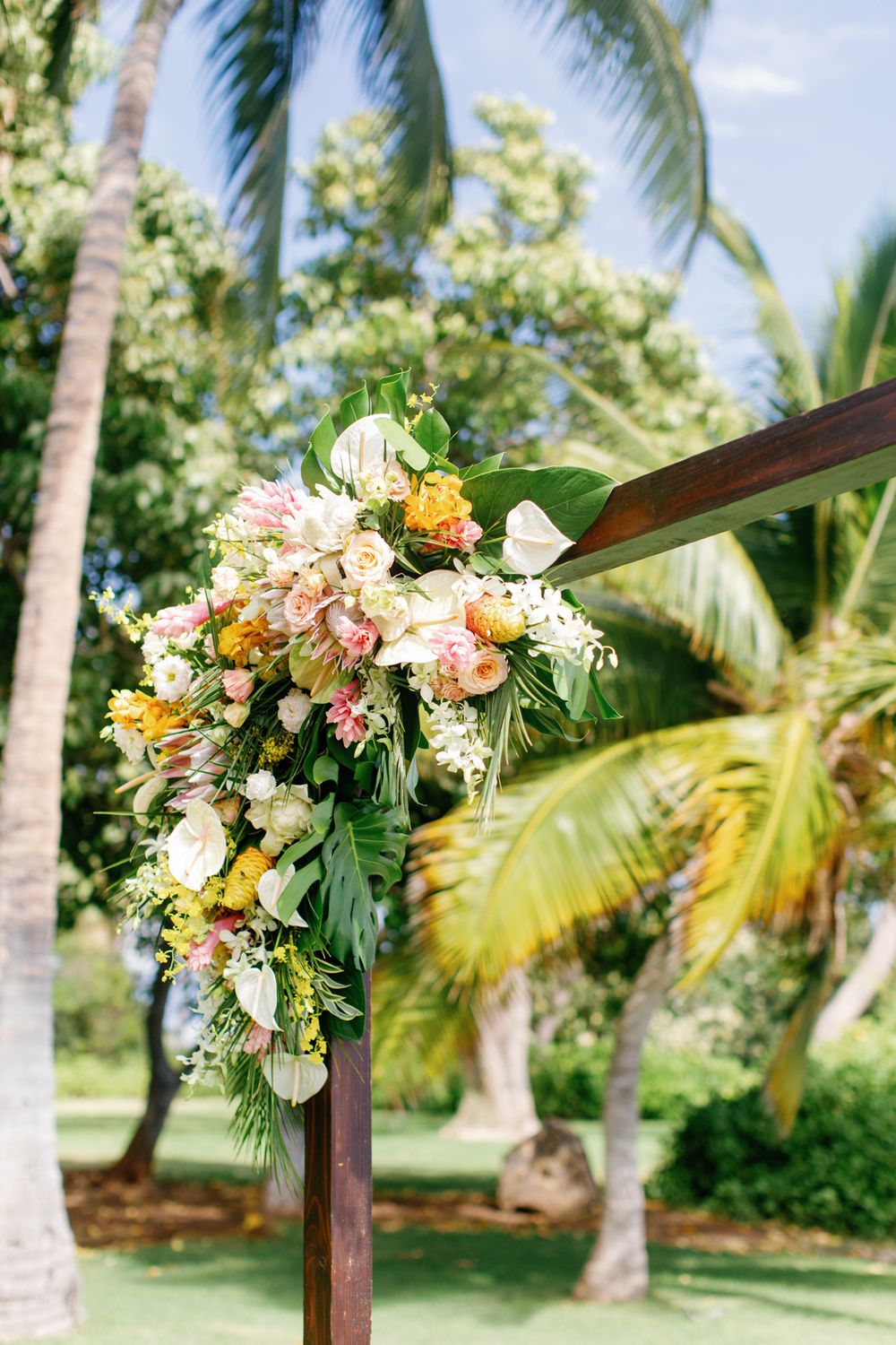 Tropical wedding arch decorated with pink and peach flowers against a backdrop of swaying palm trees in a lush garden.
