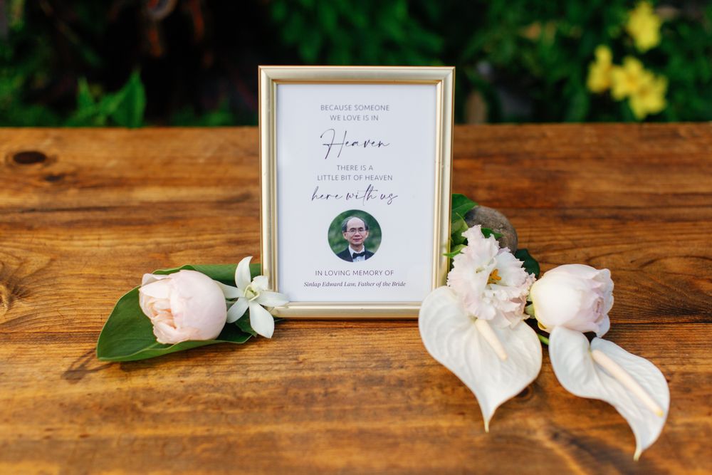 Wedding detail photo with pink roses, white anthurium leaves, and a framed table number card on wooden surface.