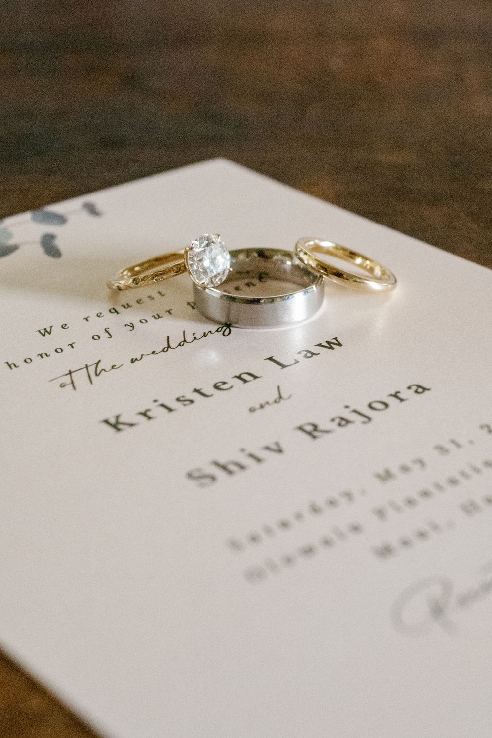 Wedding rings arranged on an elegant white invitation card showing names Kristen and Shiv resting on a wooden surface.