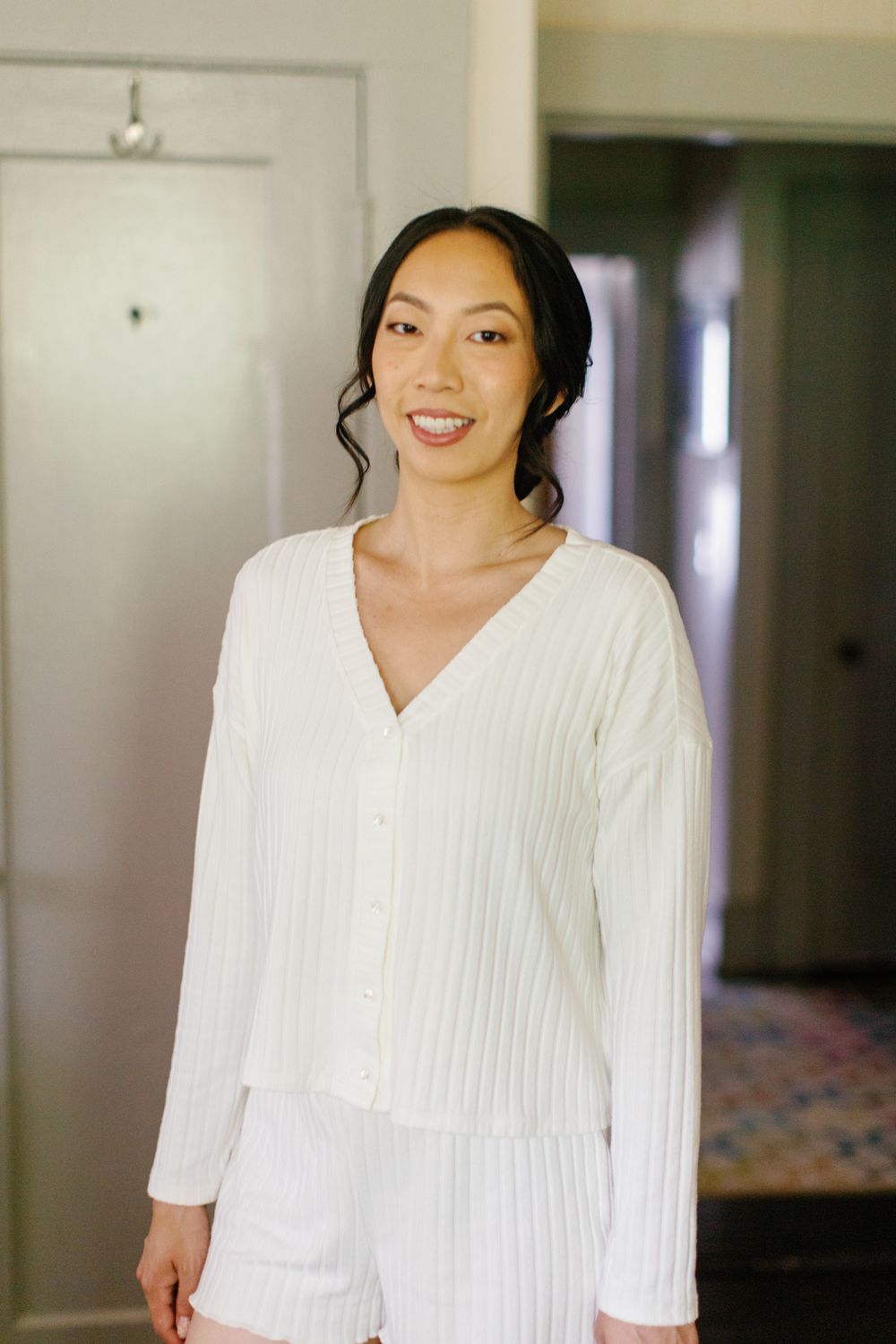 Person wearing white ribbed sweater with v-neck and long sleeves smiles warmly in interior hallway setting.