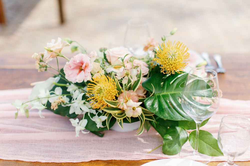 Elegant floral arrangement with yellow craspedia balls, pink roses, white jasmine flowers and monstera leaves on soft linen.