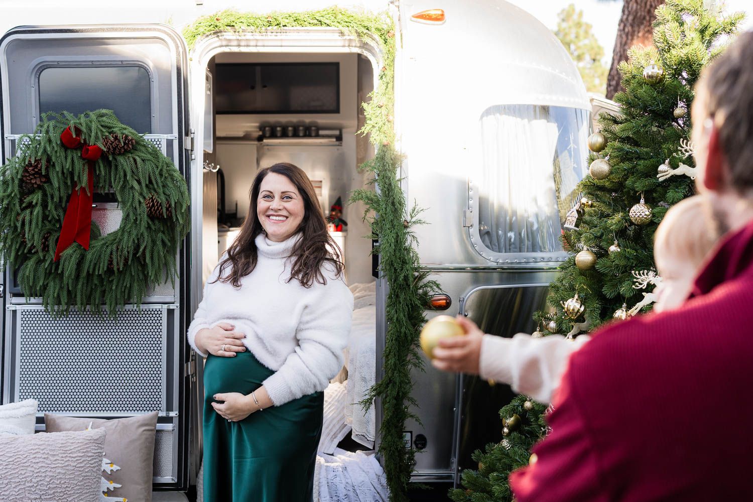 Airstream trailer decorated with holiday wreath while someone smiles from doorway as another person decorates nearby.