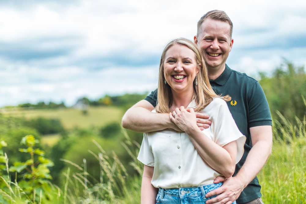 A couple laughing and embracing while standing in a green field with wildflowers and rolling hills in the background.