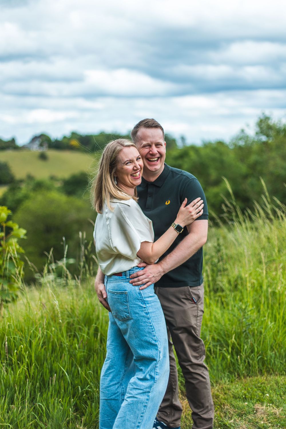 A loving couple embraces while standing in a green meadow with rolling hills and cloudy sky in the background.